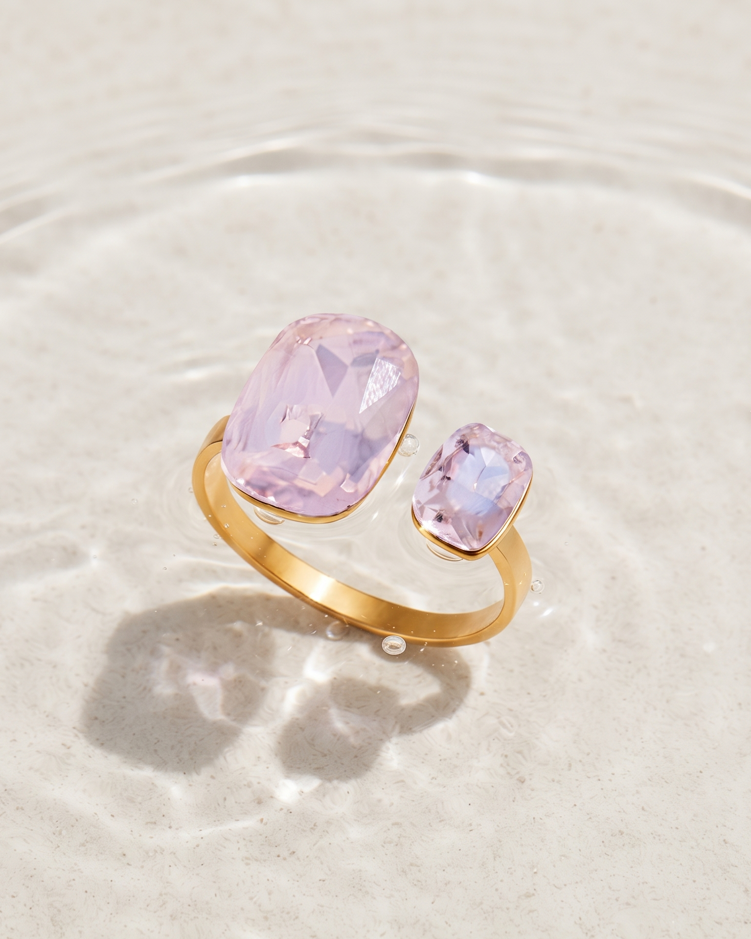 A gold ring with two lavender-pink gemstones, one large oval and one smaller rectangular, resting on a light beige textured surface with water ripples behind it.