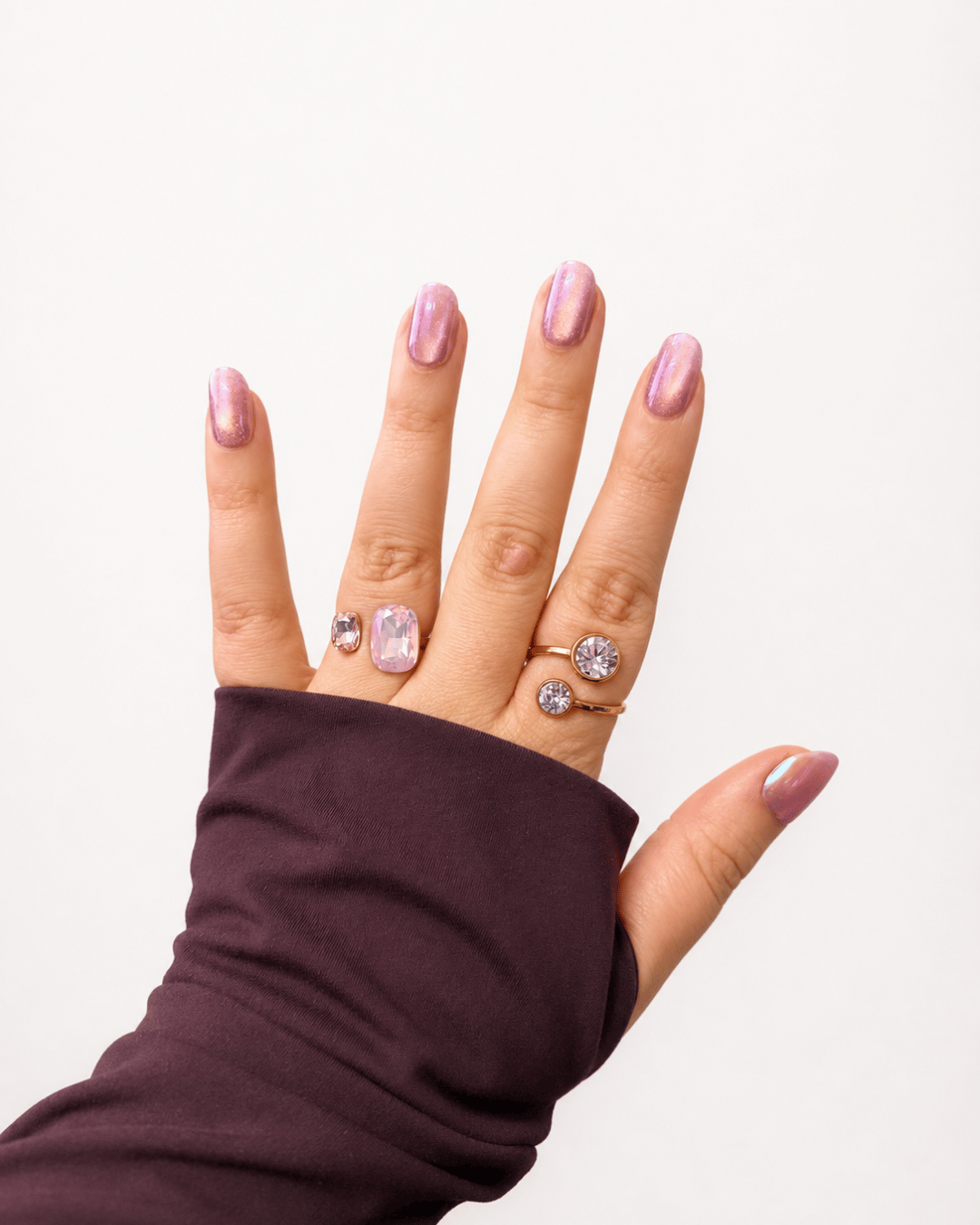 A close-up of a hand wearing three rings, including a lavender rectangular stone ring, a lavender oval stone ring, and a rose gold ring with a round clear stone. The hand has light pink manicured nails and is wearing a dark plum long-sleeve top against a plain white background.