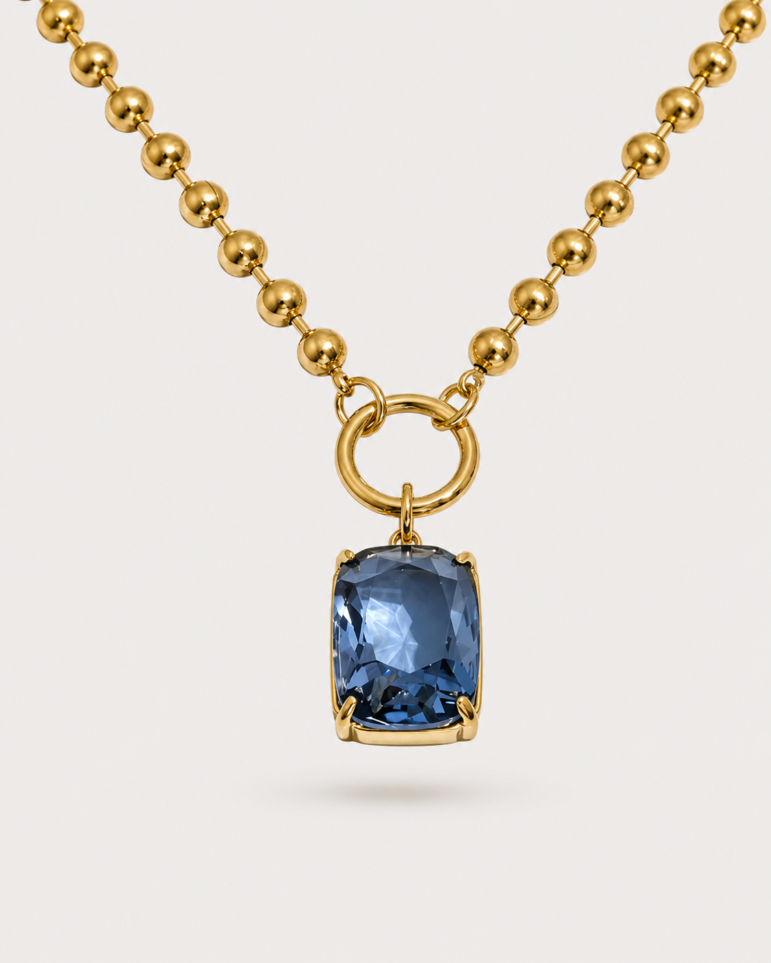 A gold ball-chain necklace featuring a rectangular cushion-cut blue gemstone pendant in a gold bezel setting, suspended from a gold ring connector on a light gray background.