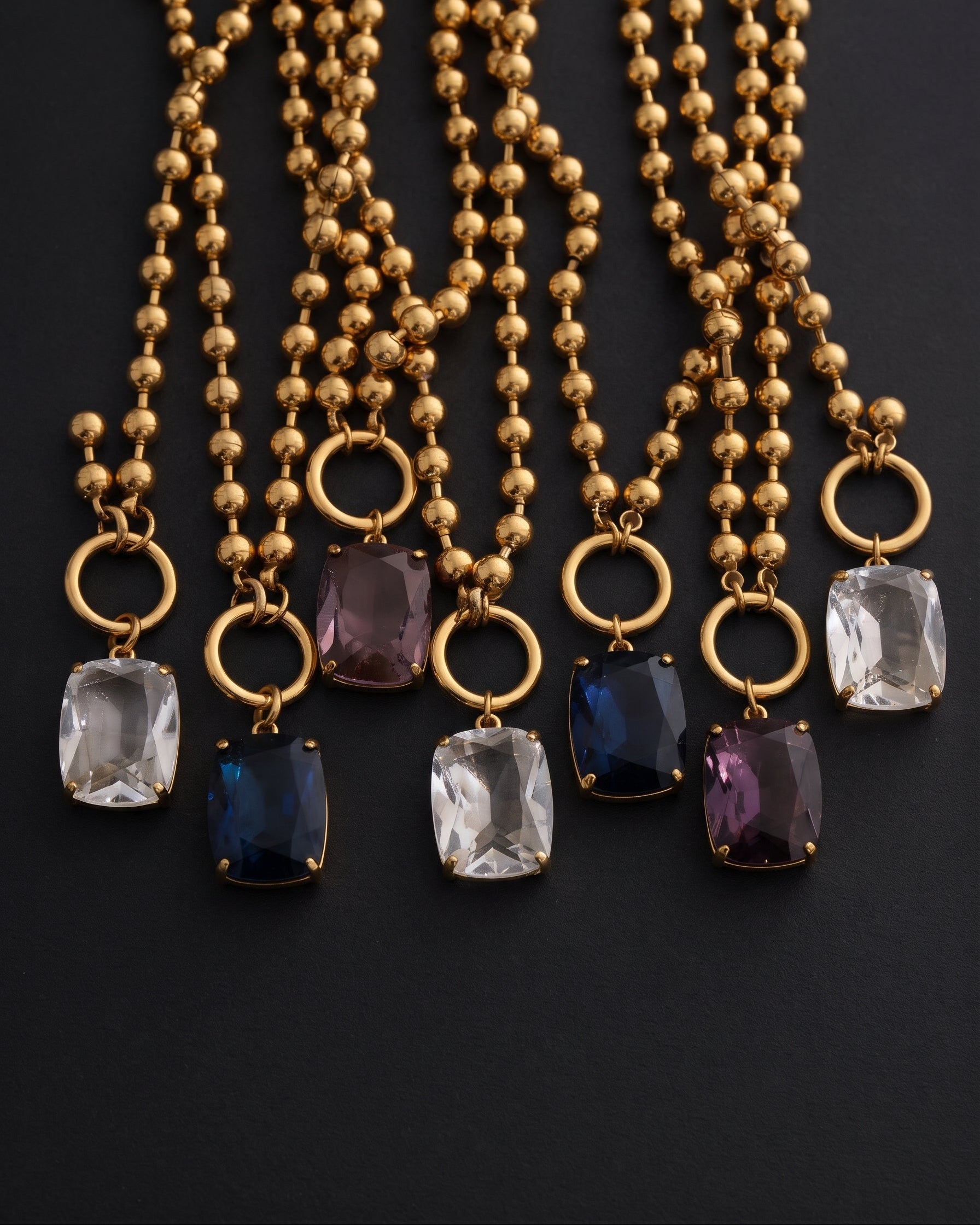 Six gold beaded necklaces with round ring pendants and rectangular gemstone charms in clear, light blue, and purple, arranged on a dark background.