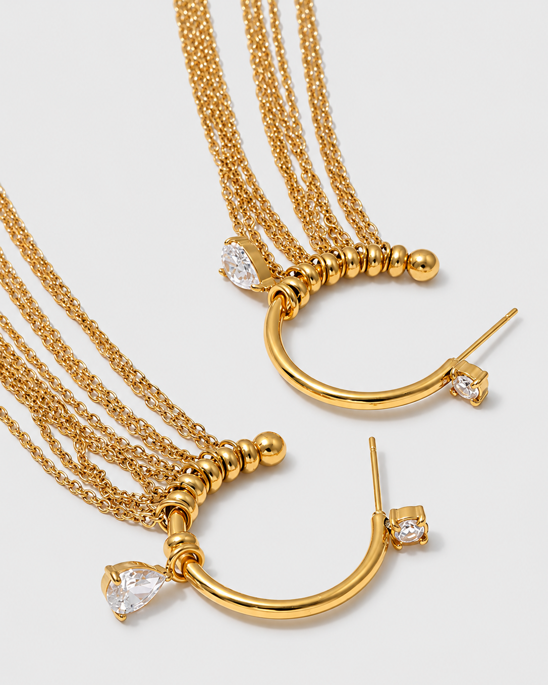 A pair of gold crescent-shaped hoop earrings with multiple layered chains, small bead details, and clear teardrop and round crystals, shown on a light gray background.