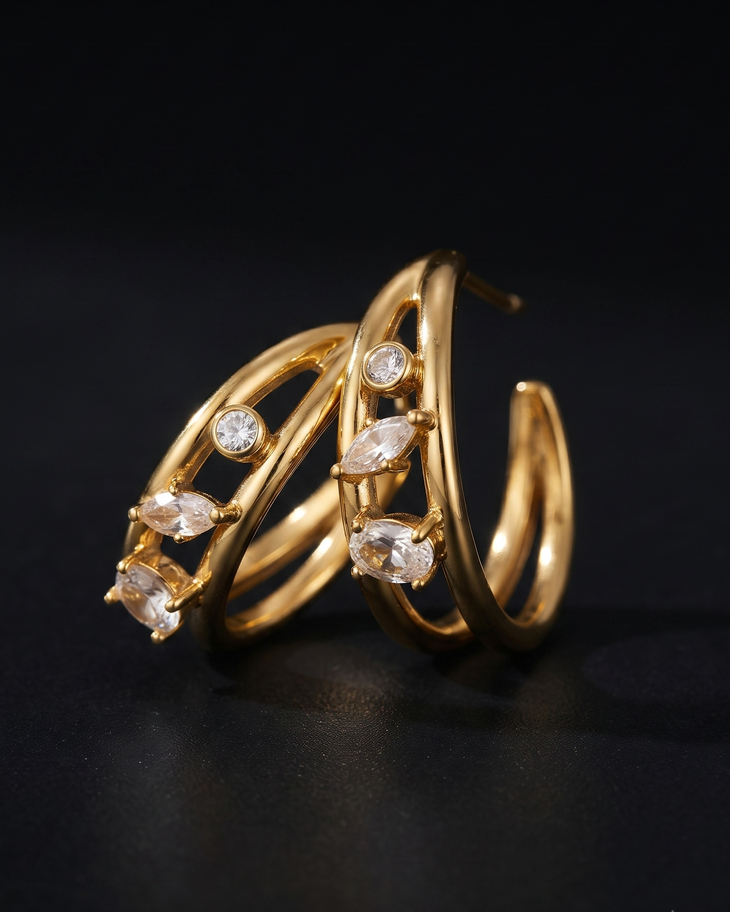Gold hoop earrings with marquise-cut clear crystals, displayed on a dark reflective surface.
