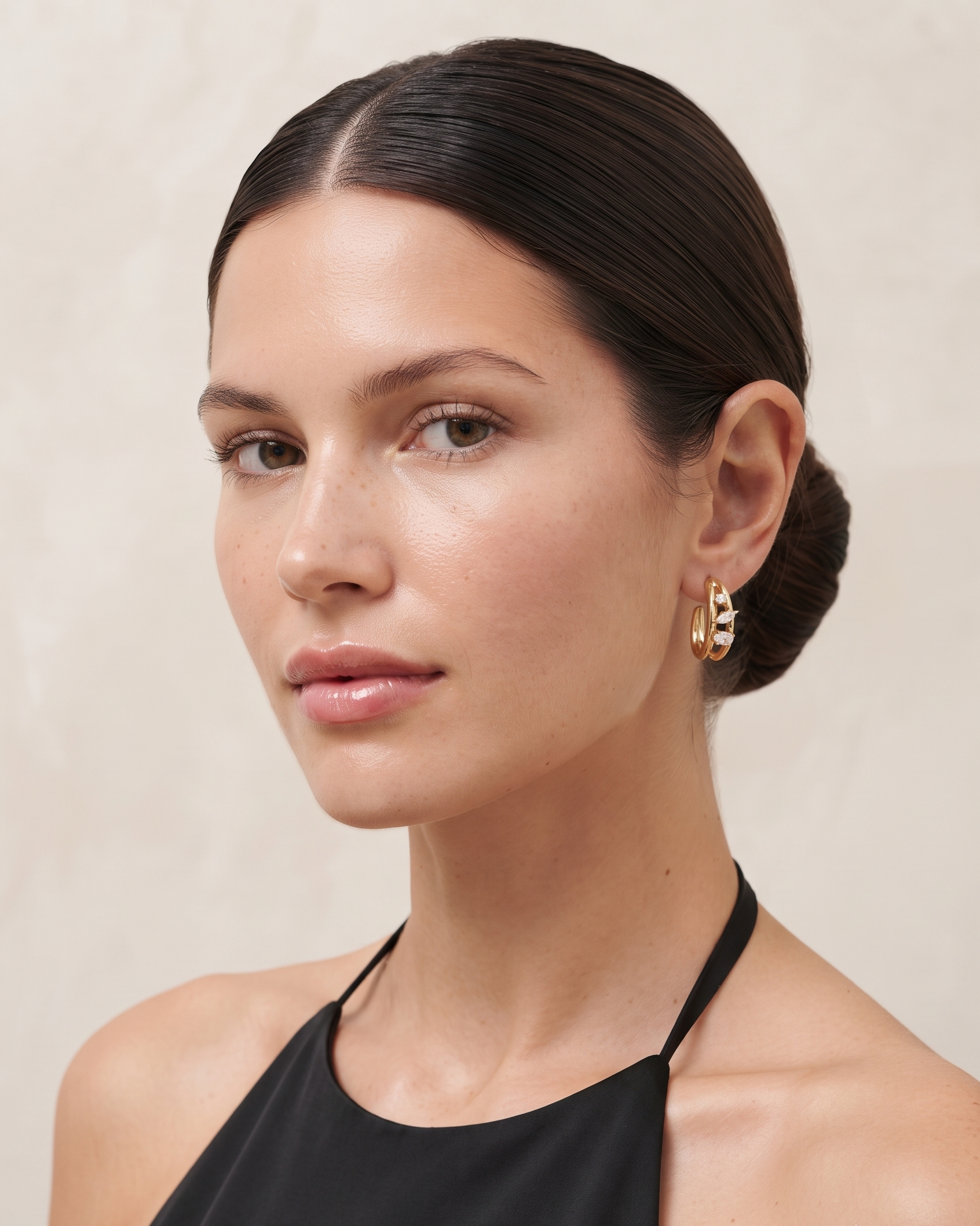 A close-up portrait of a woman with dark hair in a low bun wearing a black halter top and a small gold hoop earring with a textured, organic gold design.