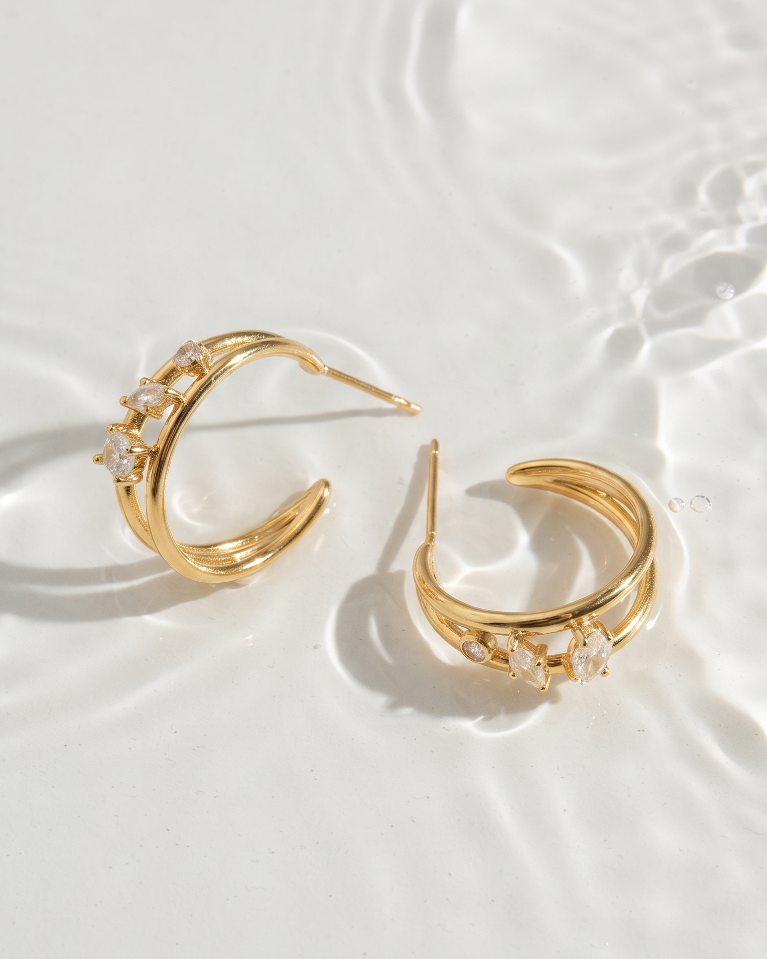A pair of gold crescent-shaped hoop earrings with clear crystal accents, displayed on a white surface with water ripples.