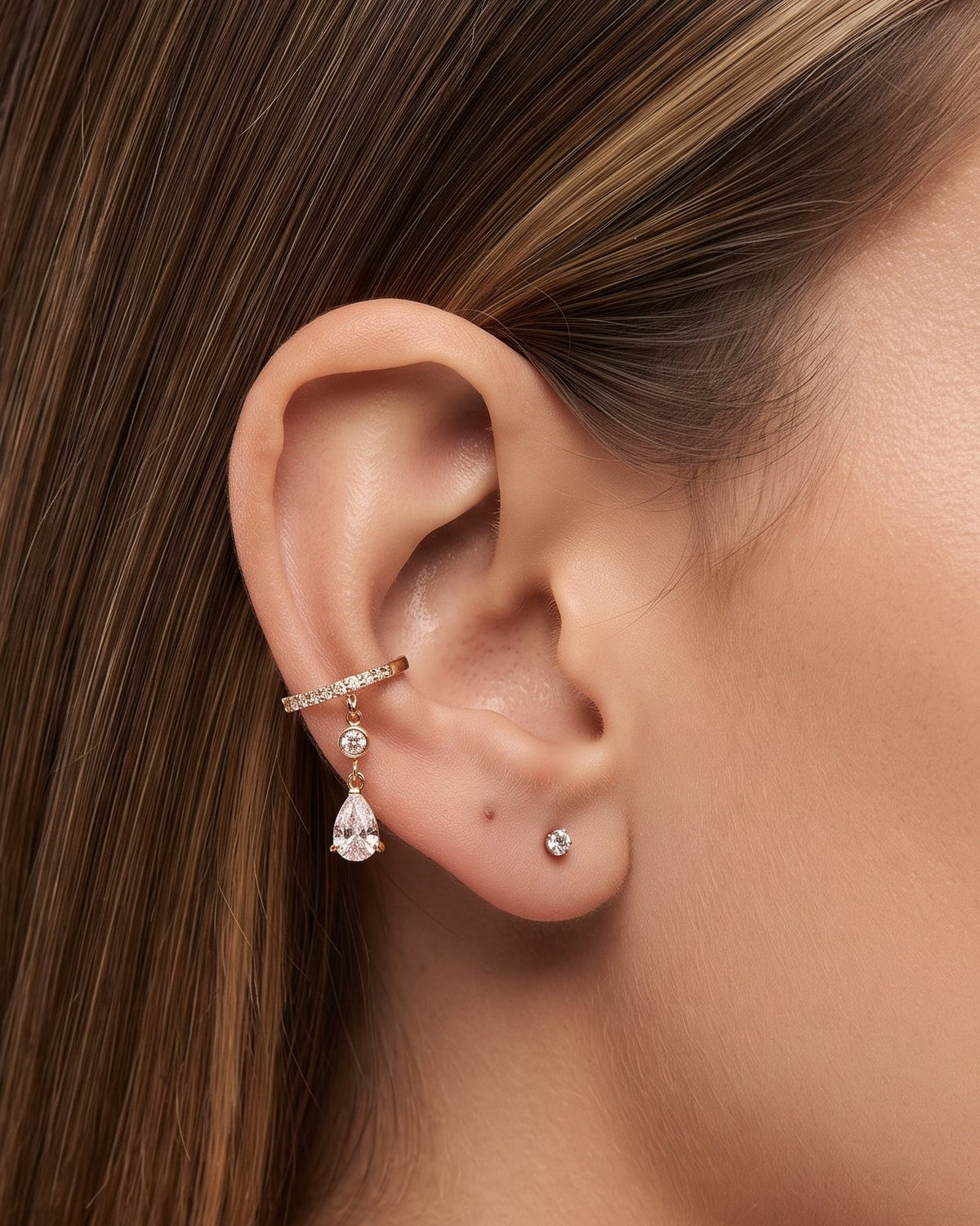 A close-up of a model's ear wearing a rose gold ear cuff with a dangling teardrop crystal charm, alongside a small stud earring.