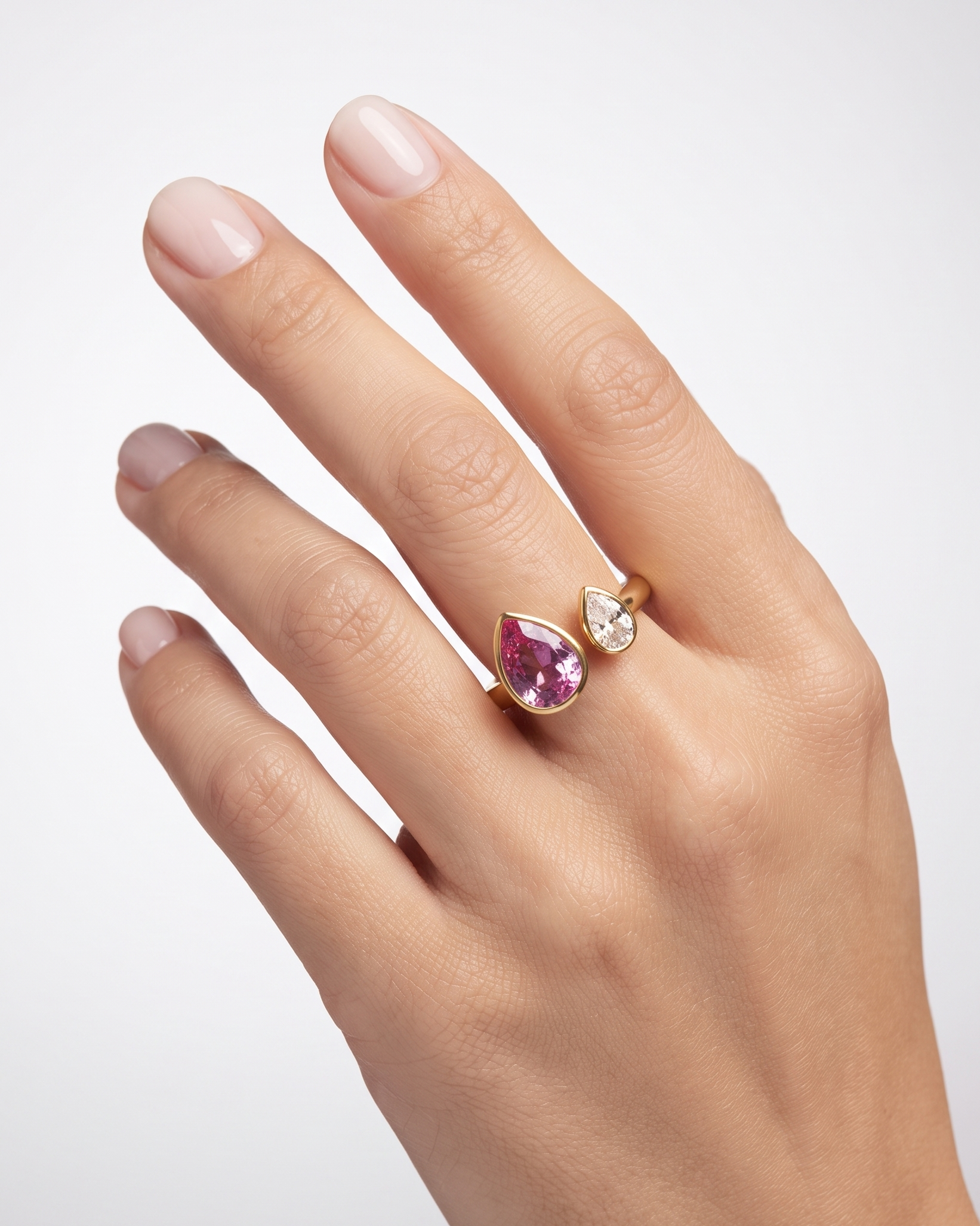 A close-up of a hand wearing a gold ring with a teardrop-shaped pink gemstone and a smaller clear teardrop gemstone beside it, set against a light gray background.