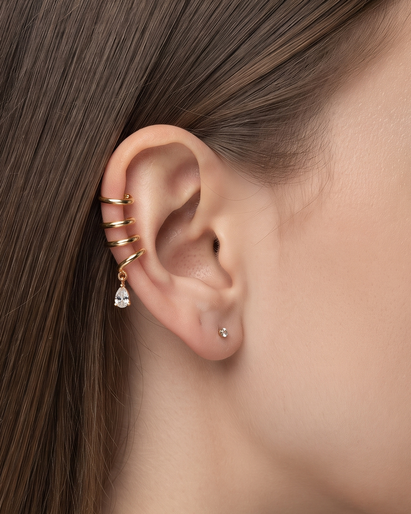 A close-up side profile of a woman's ear wearing a stack of gold ear cuffs, including a small dangling clear crystal charm, against a soft gray background.