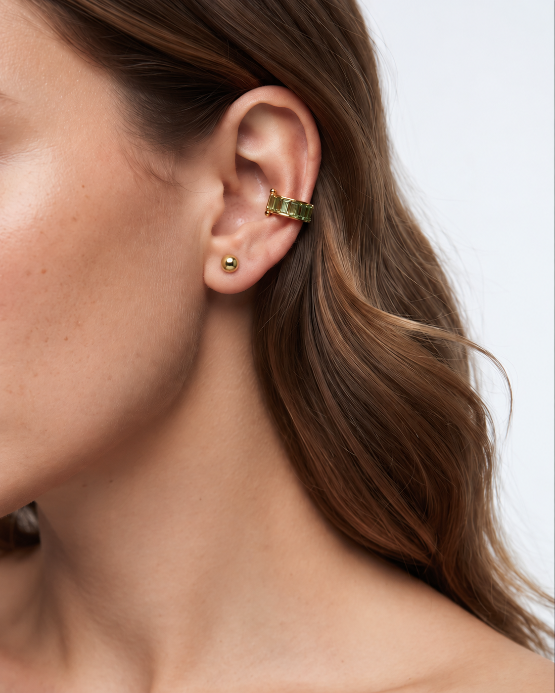 A close-up side profile of a woman's ear wearing a slim gold ear cuff with small clear stones, against a light gray background.