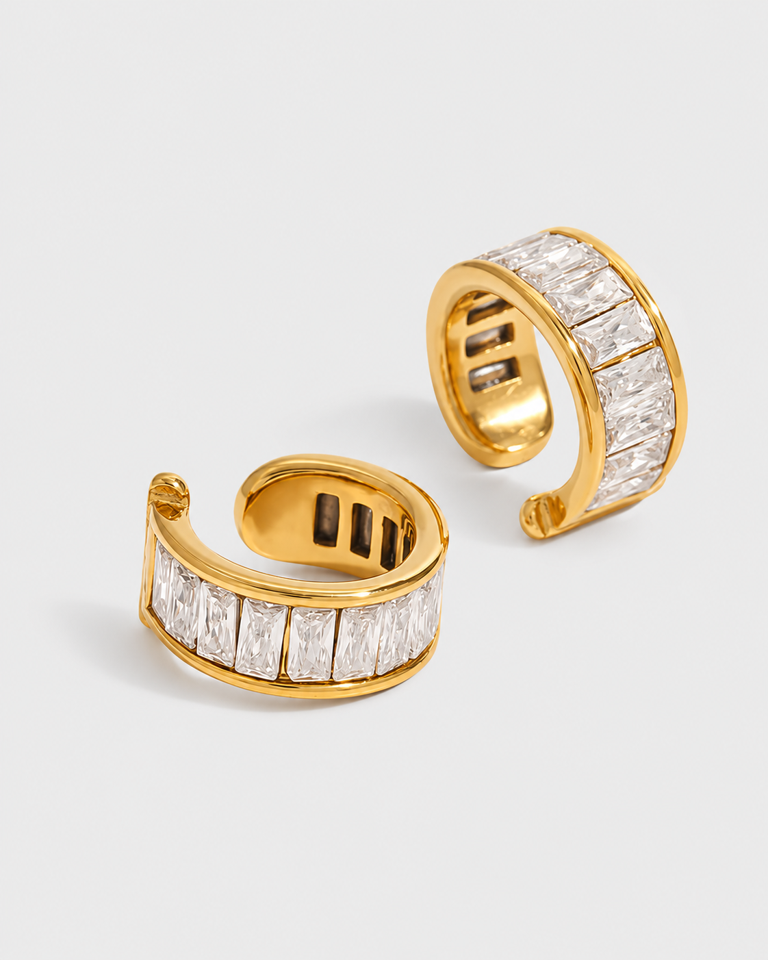 A pair of gold huggie hoop earrings paved with baguette-cut clear stones, displayed on a light gray background.