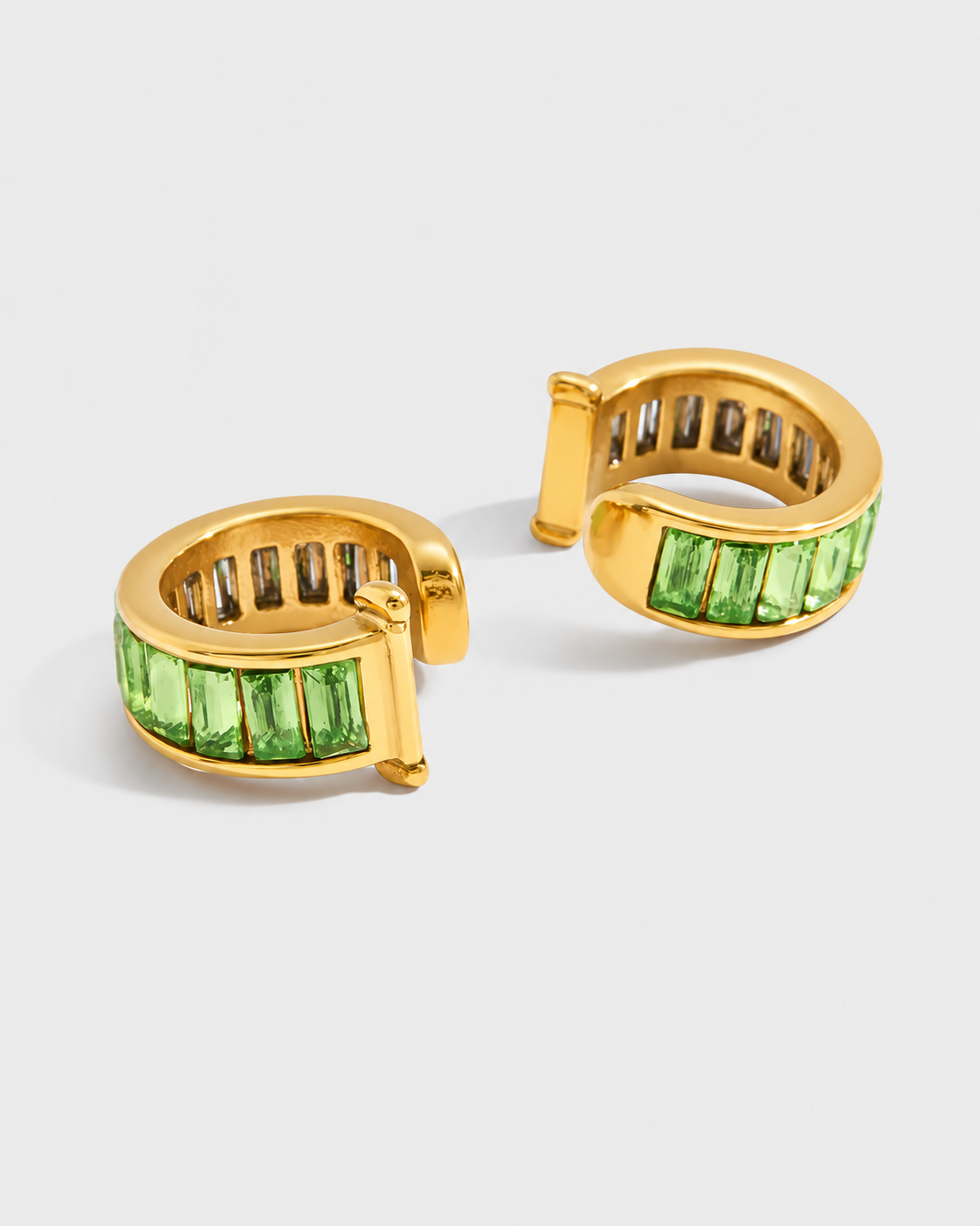 A pair of gold huggie hoop earrings embellished with baguette-cut green gemstones, displayed on a light gray background.