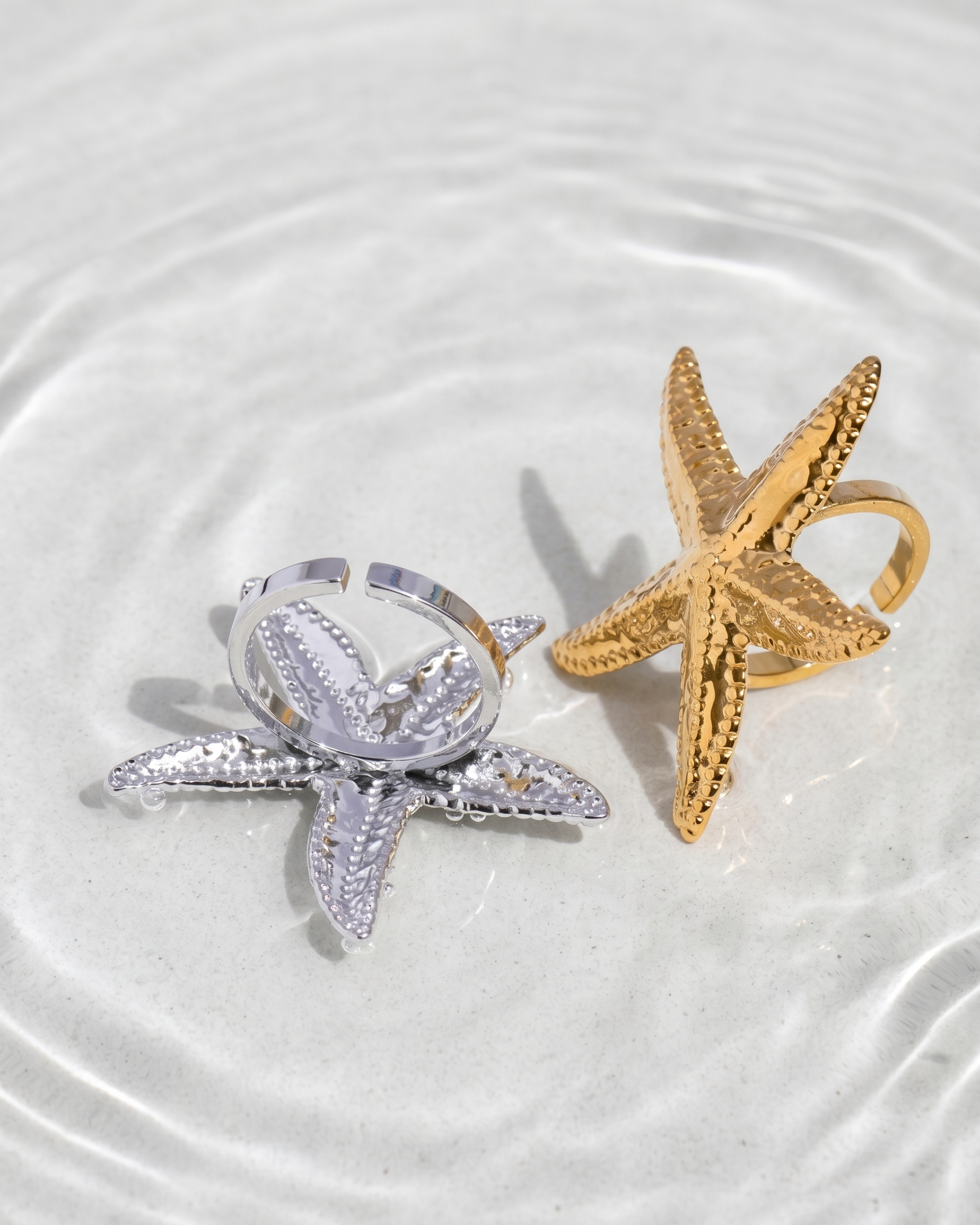 Two starfish-shaped rings, one silver and one gold, on a white surface with water ripples.