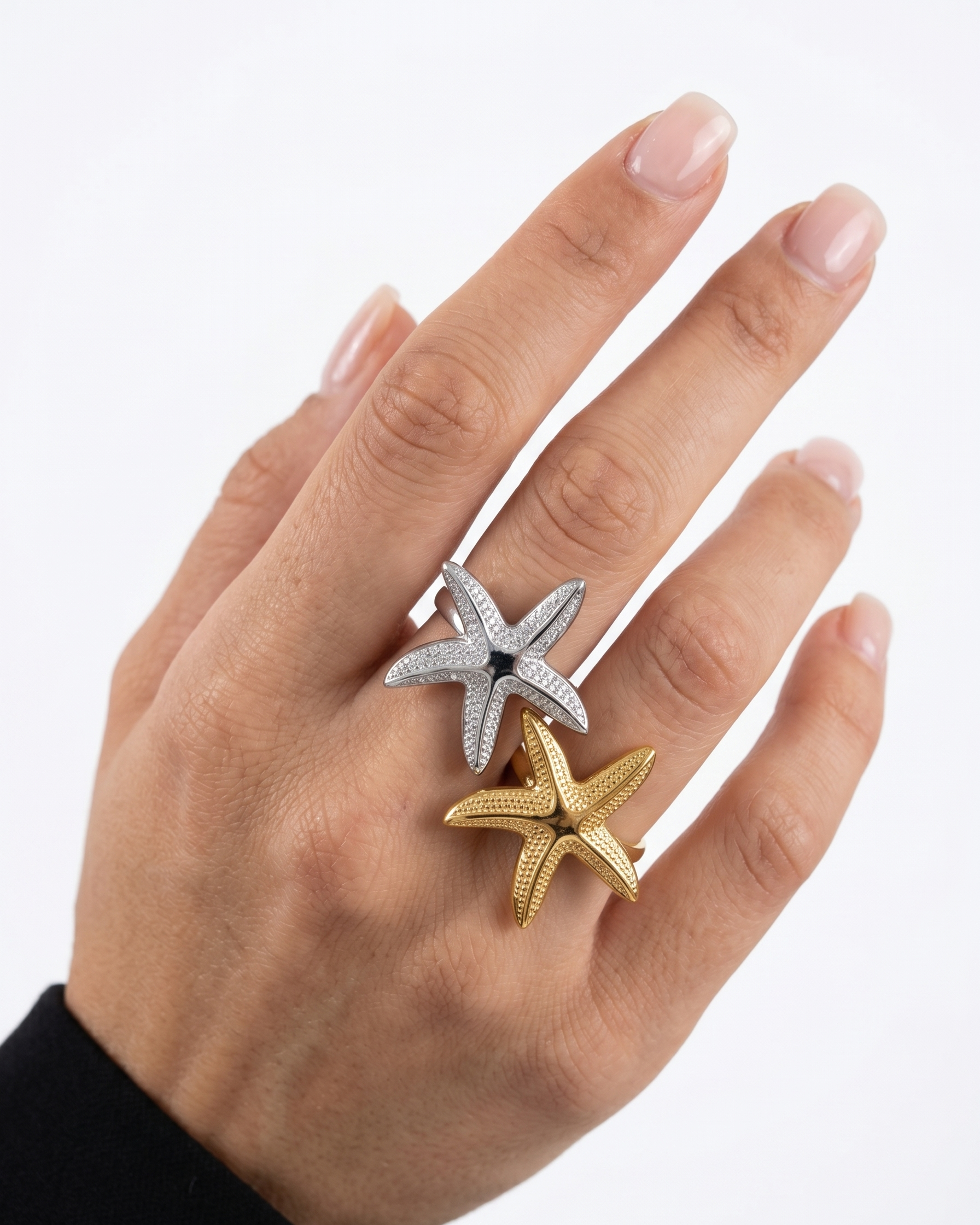 Hand wearing two starfish-shaped rings, one gold and one silver, on a white background.