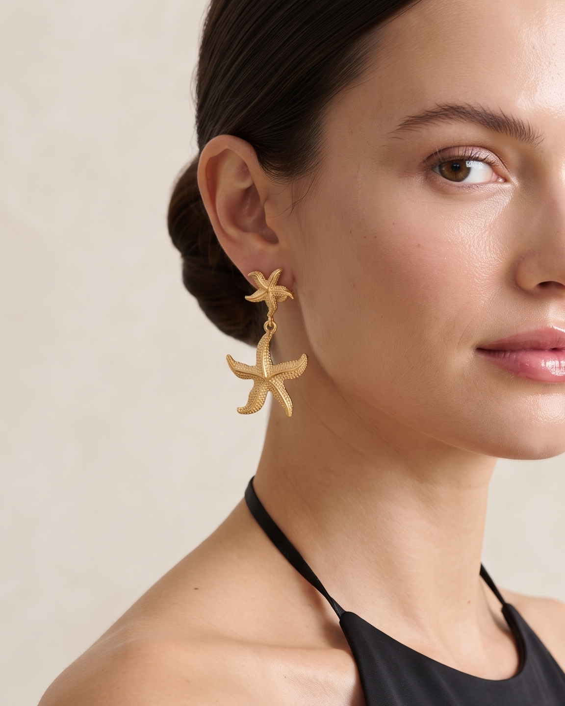 A woman with her hair pulled back wears a pair of dangling starfish earrings in a warm gold tone, paired with a black halter top against a soft beige background.