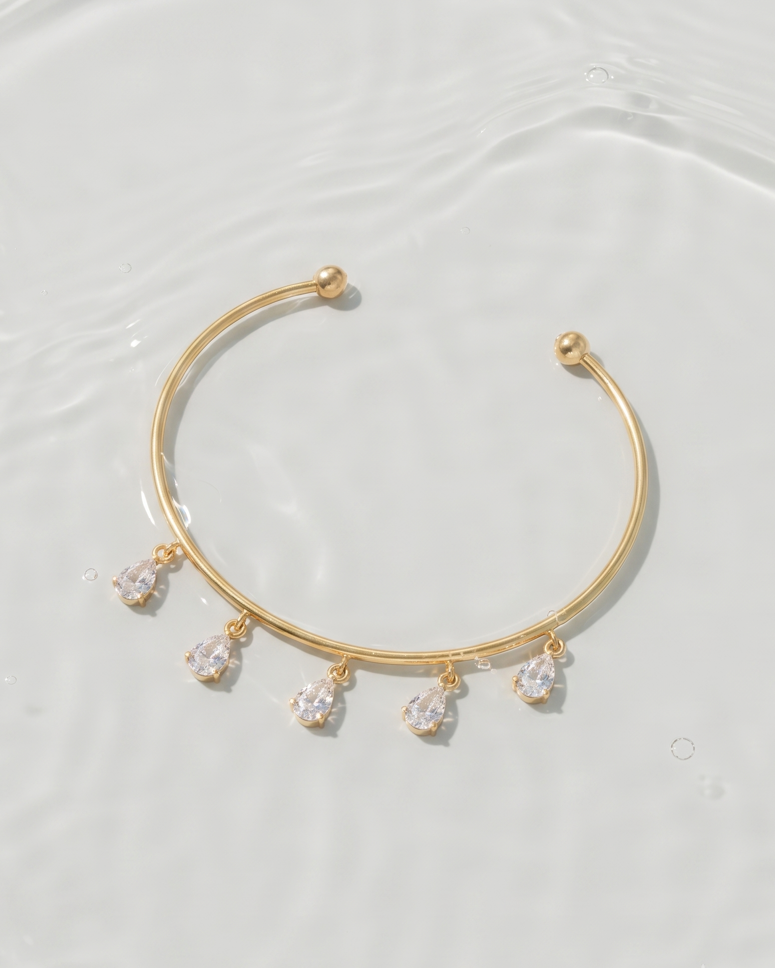 A delicate gold cuff bracelet with five dangling clear teardrop gemstones, displayed on a light, water-textured surface.