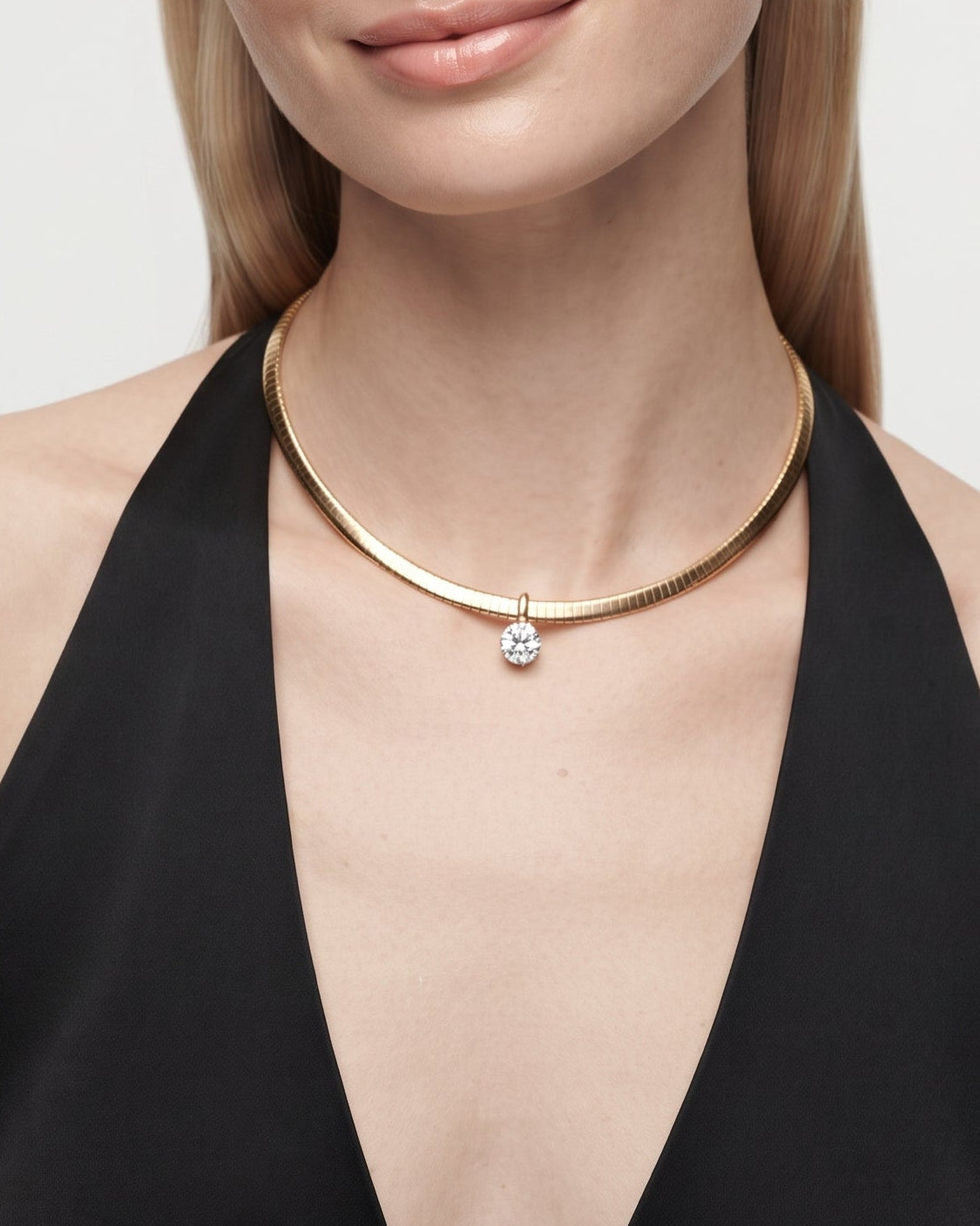 A blonde model wearing a slim gold collar necklace with a small clear gemstone pendant, styled with a black halter top against a light gray background.