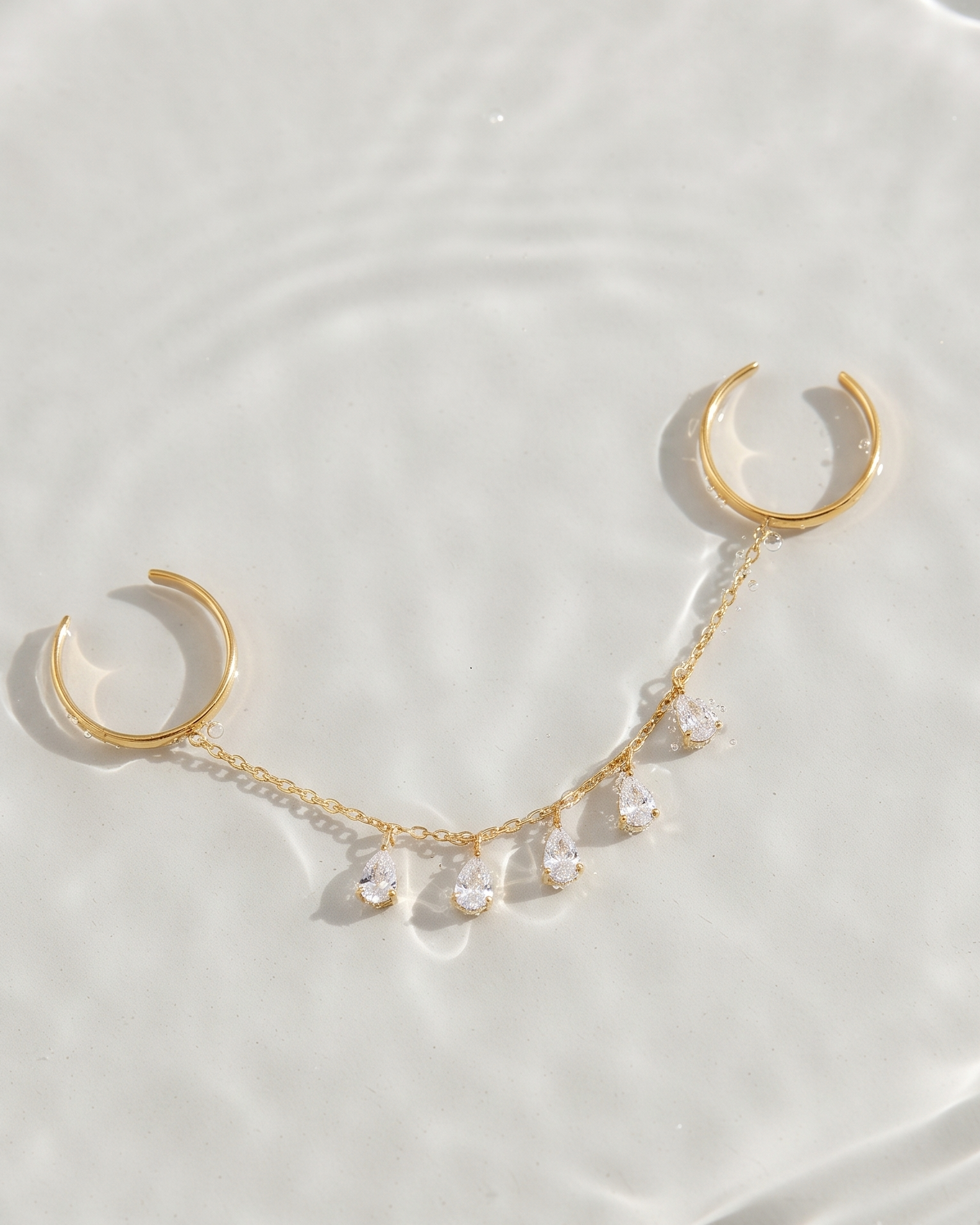 Gold-toned body jewelry featuring two open-ended crescent hoop ends connected by a delicate chain with small clear rhinestone drops, arranged on a glossy white surface with soft reflections.