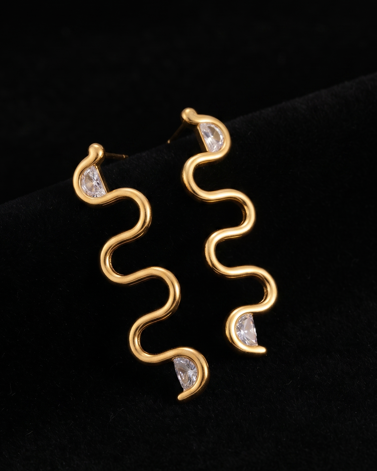 A pair of gold wavy spiral drop earrings with small clear gemstone accents, displayed on a black velvet surface.