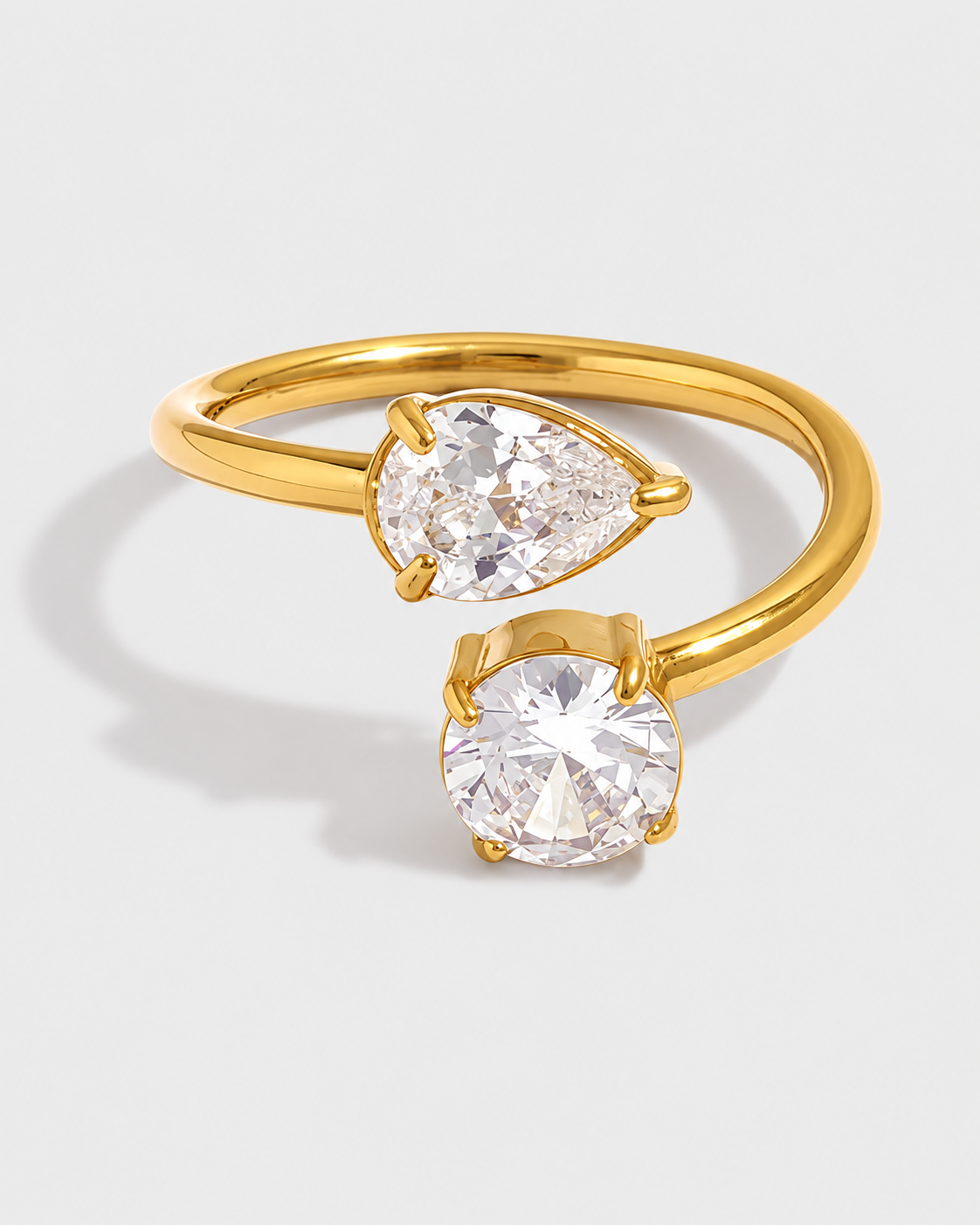 A gold open-band ring featuring two clear gemstones, one pear-shaped and one round, set against a light gray background.