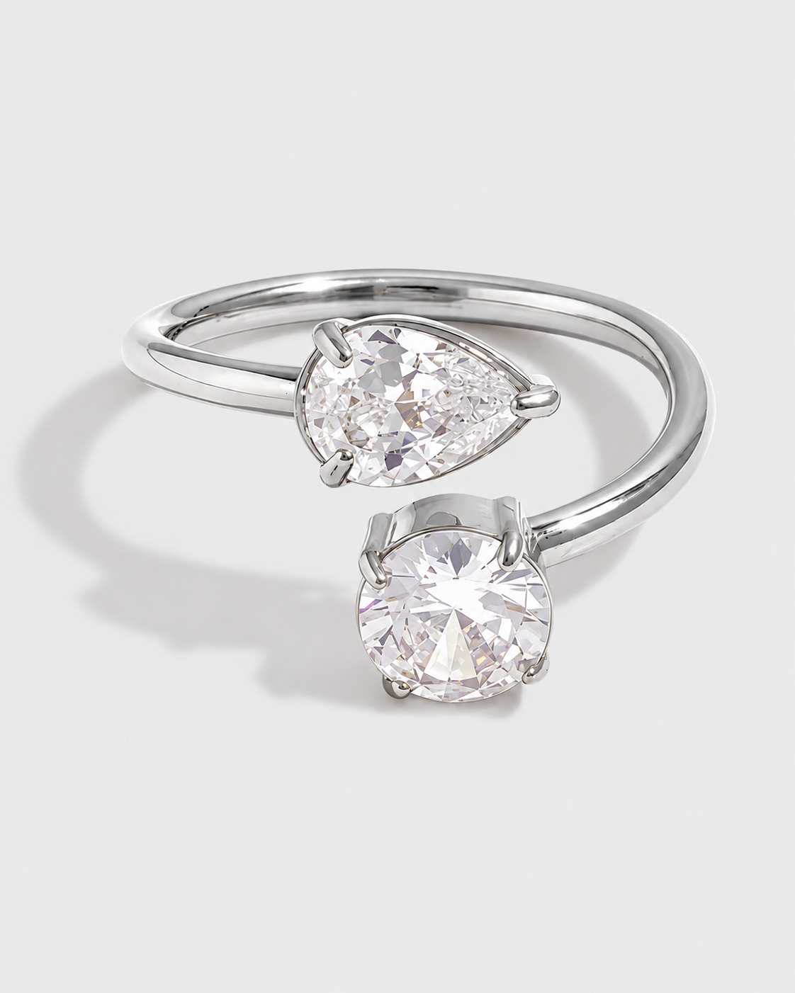 A silver-toned open band ring featuring two clear gemstones, one pear-cut and one round-cut, set against a light gray background.