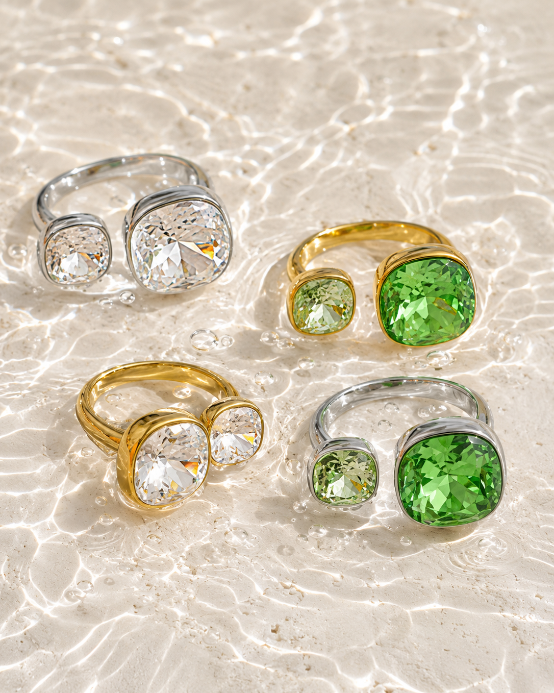Four rings with large round clear and green gemstones in silver and gold settings, arranged on a sandy beach with shallow water ripples.