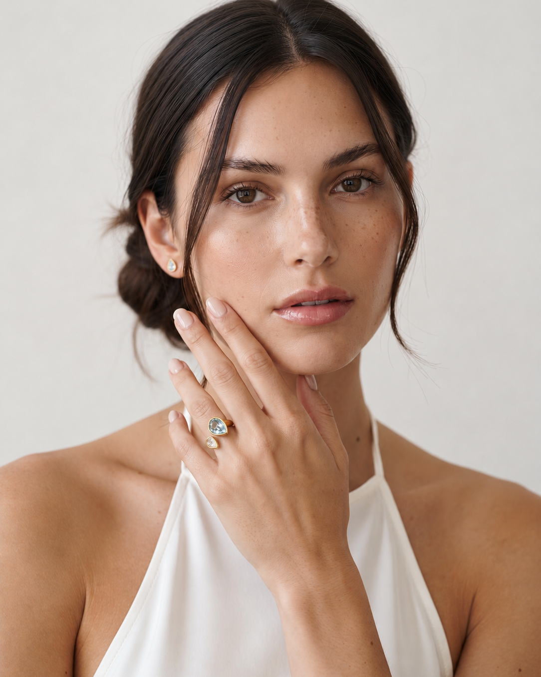 A close-up portrait of a woman wearing a delicate gold ring with a small clear gemstone on her finger, set against a soft, light gray background.
