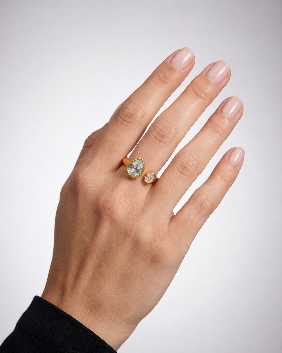 A close-up of a hand wearing two gold rings, with the larger ring featuring a pear-cut light blue stone and the smaller ring featuring a round clear stone, set against a neutral gray background.