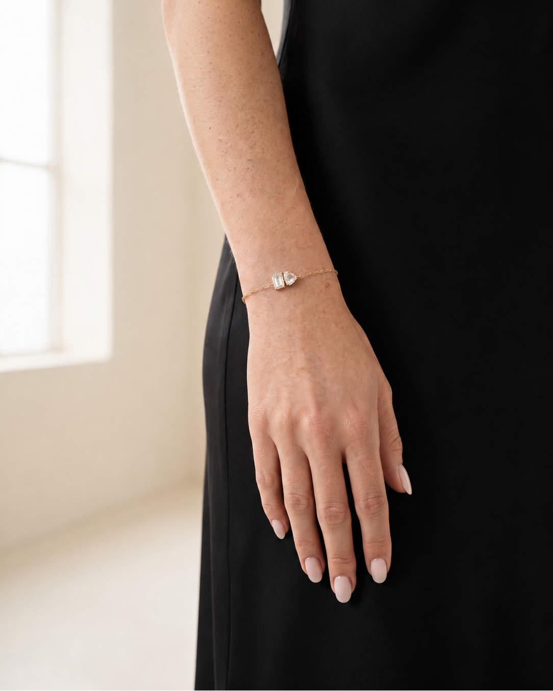 A delicate gold chain bracelet with a small heart-shaped crystal pendant, shown worn on a woman's wrist. The model is wearing a black dress and standing in a brightly lit room.