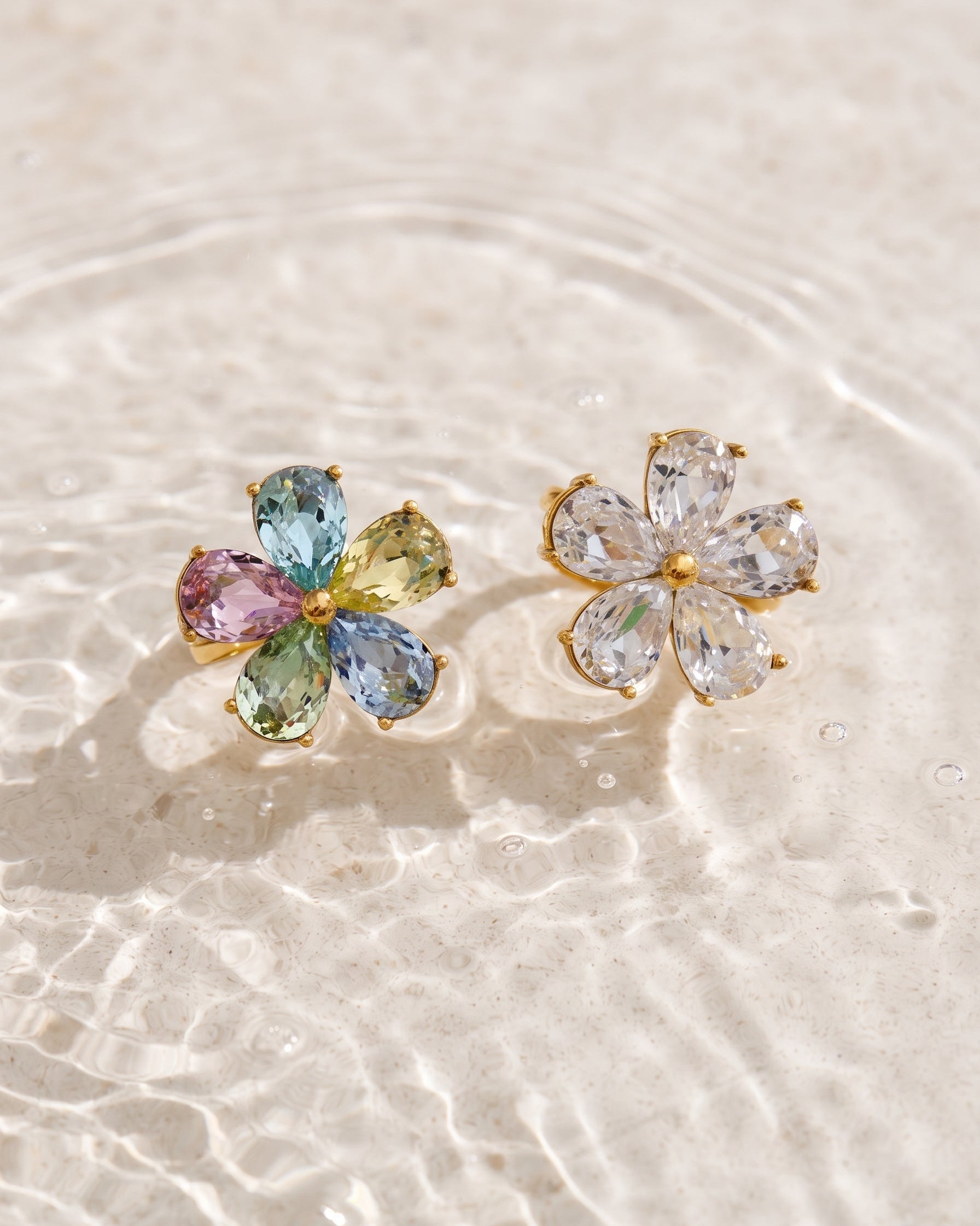 A pair of gold flower-shaped stud earrings resting on shallow water. One earring features multicolored pastel gemstones (blue, green, pink, yellow), while the other features clear, colorless gemstones.