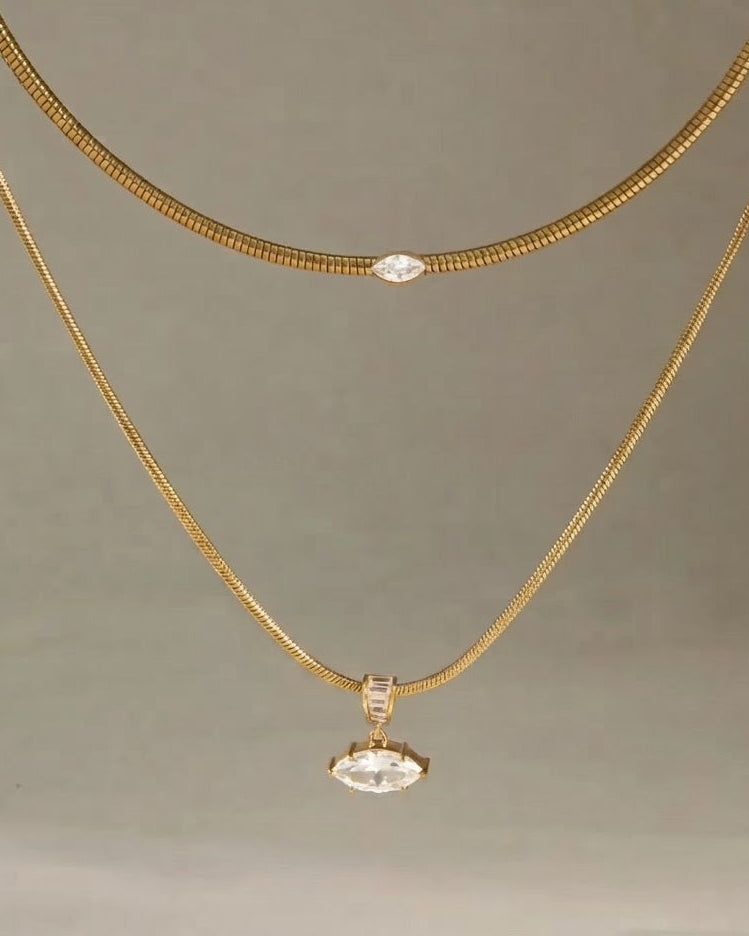 A gold necklace set featuring a textured snake chain and a fine chain with a marquise-cut clear gemstone pendant, displayed against a neutral gray background.