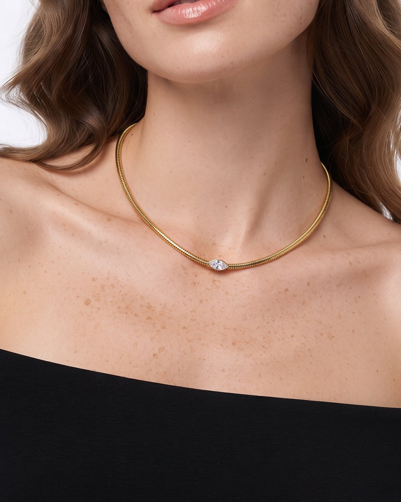 A close-up of a woman wearing a delicate gold snake-chain necklace with a small central clear gemstone, paired with a black off-the-shoulder top against a light gray background.