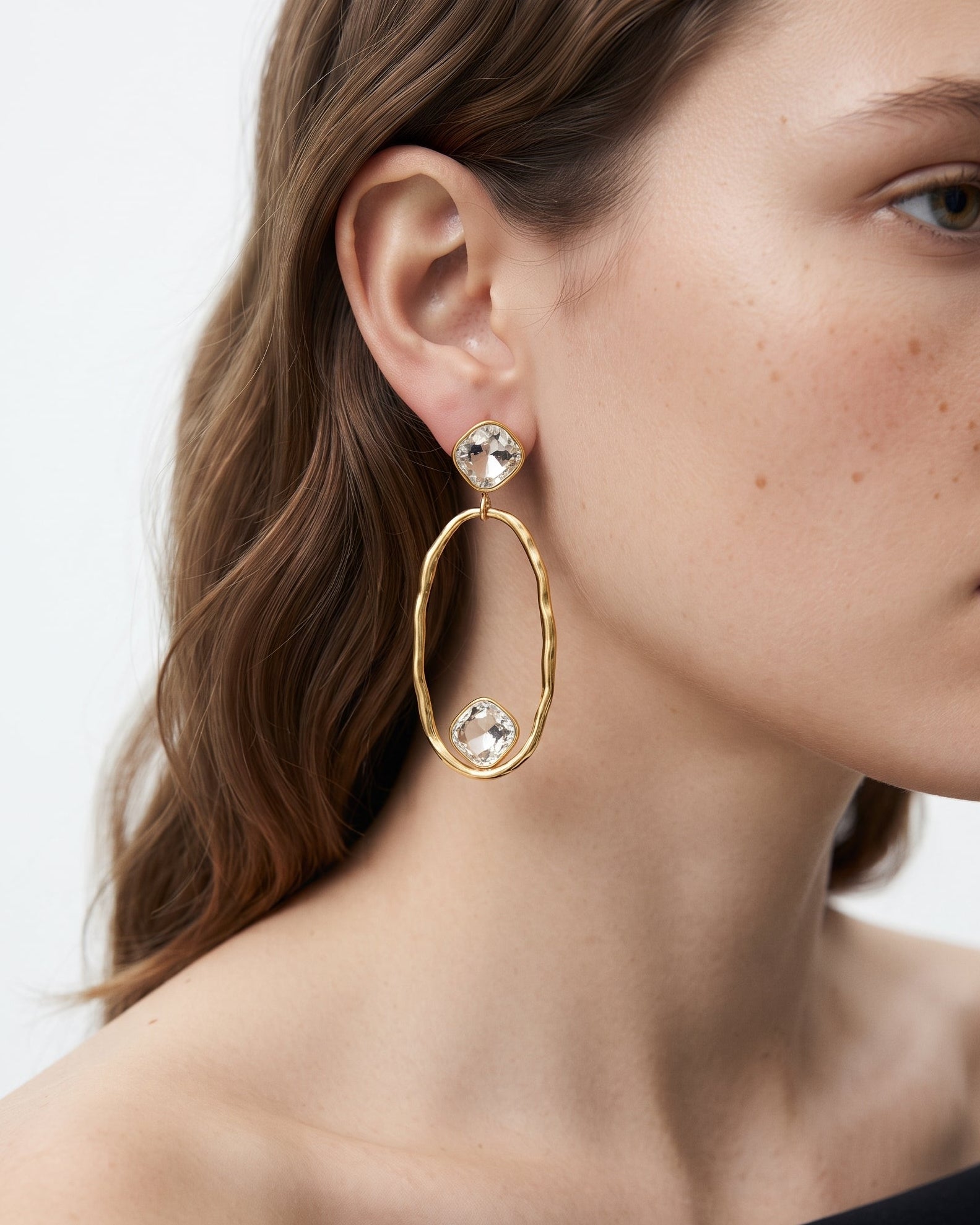 A close-up of a woman wearing a gold dangle earring with a large twisted oval hoop, a clear faceted crystal stud, and a smaller clear crystal accent.