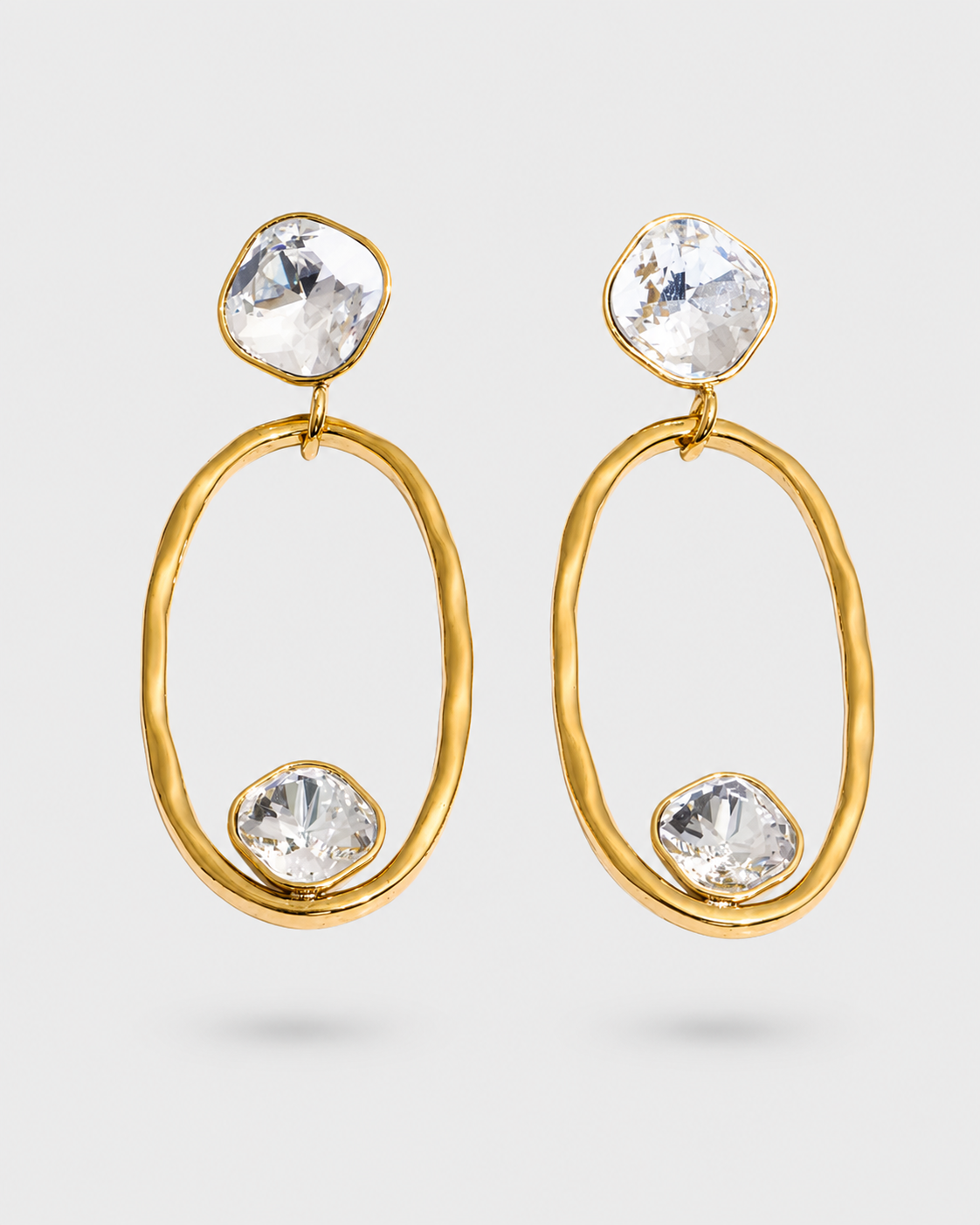 A pair of gold drop earrings featuring organic-shaped oval hoops, clear faceted gemstones at the top and bottom, and a small gold connector at the top, shown against a light gray background.