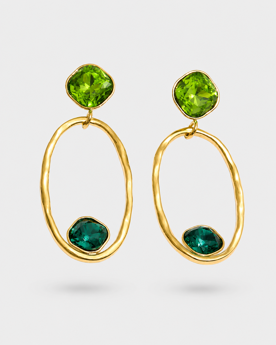 A pair of gold drop earrings featuring a large textured oval frame, a faceted green top stone, and a smaller green bottom stone, shown on a light gray background.