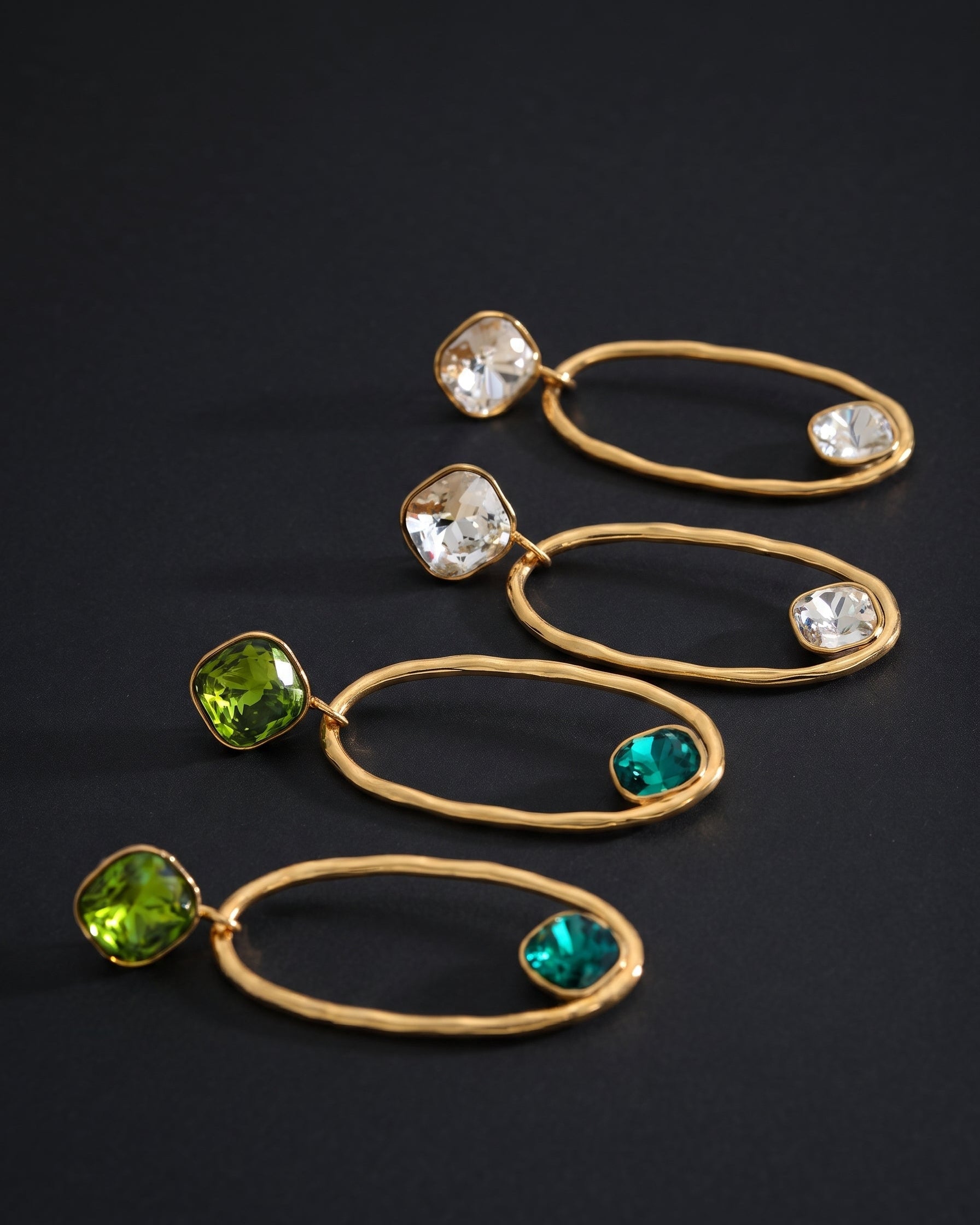 Four gold hoop dangle earrings arranged diagonally on a dark background. Each earring features a twisted gold oval hoop with a bezel-set clear faceted stone on one end and a bezel-set green stone in one of two shades on the other.