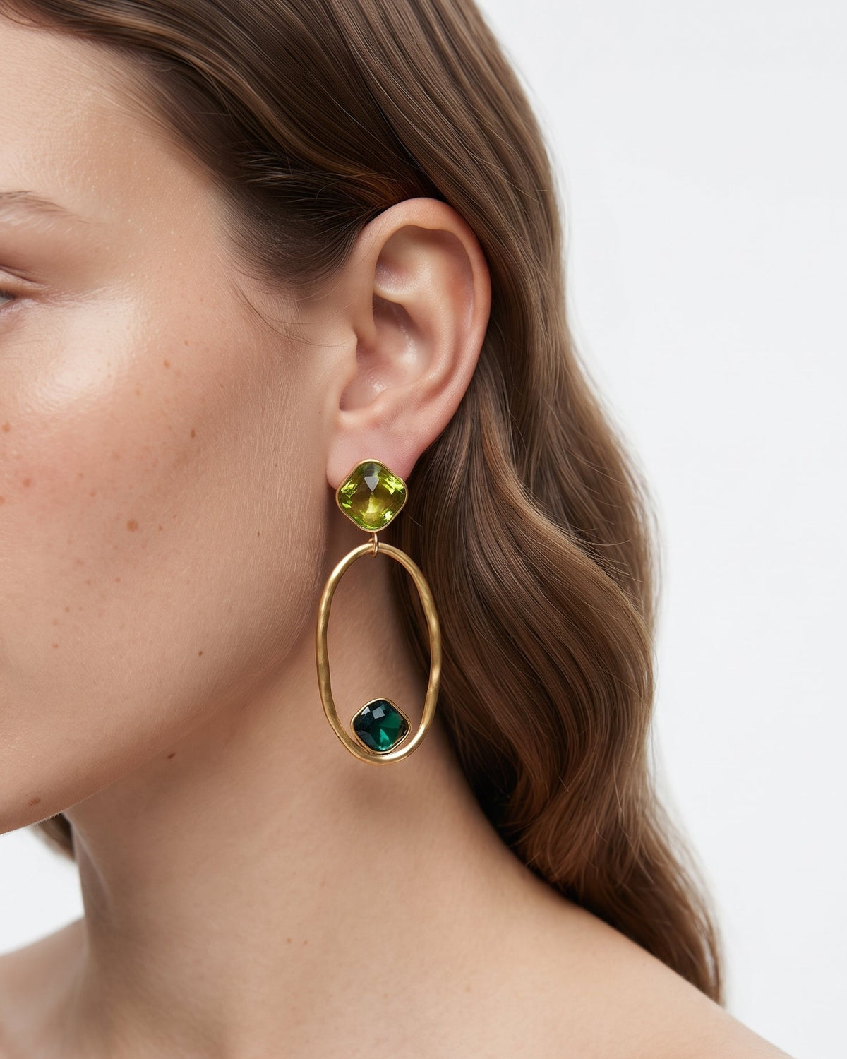 A close-up of a woman's ear wearing a gold dangle earring with a green faceted gemstone at the top and a larger oval gold hoop featuring a small dark green gemstone near the bottom.