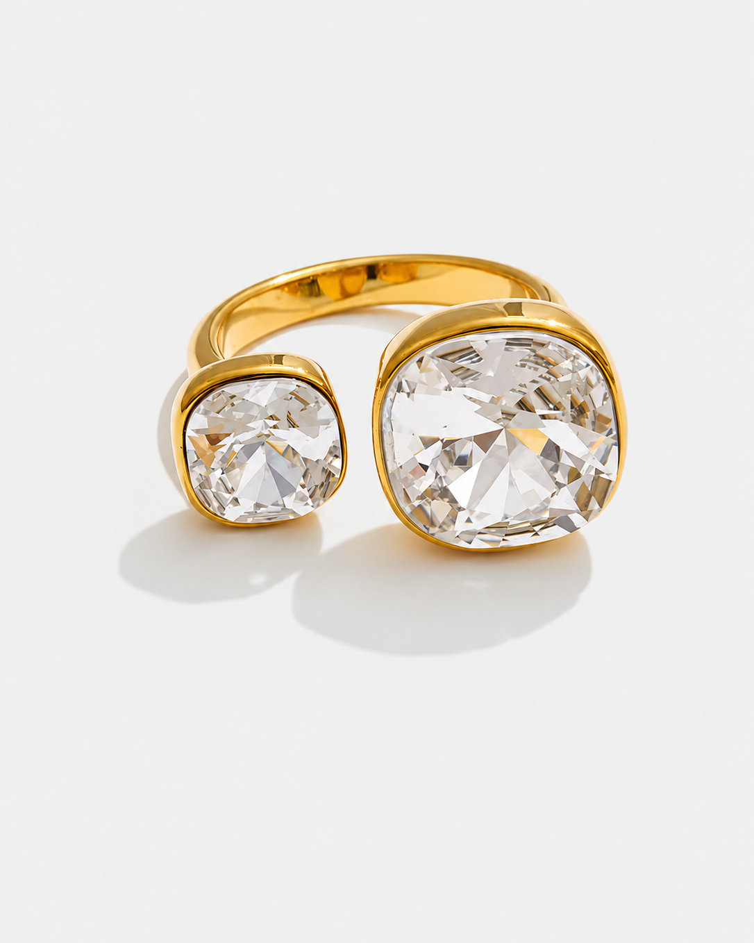 A gold ring with two cushion-cut clear gemstones in bezel settings, displayed on a light gray background.