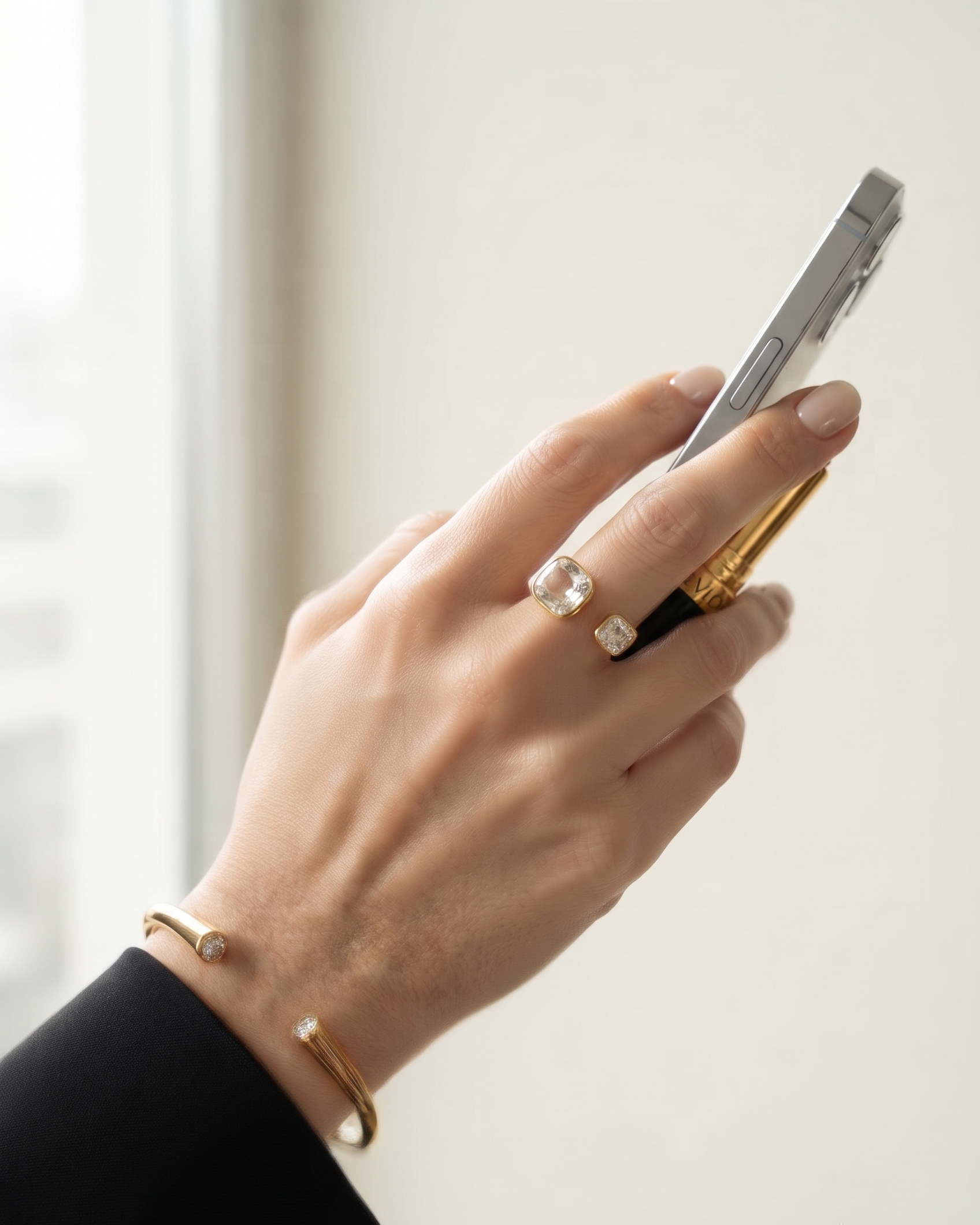 A close-up of a person's hand holding a silver smartphone, wearing a gold open cuff bracelet and a gold ring with a clear gemstone.