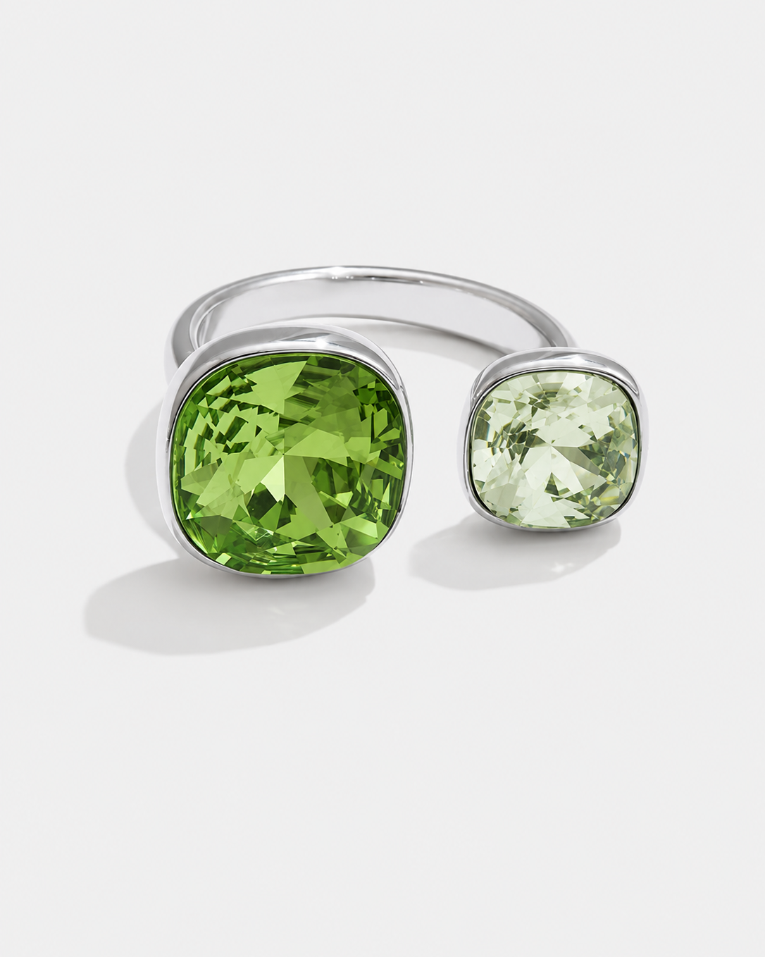 A silver-toned open ring band featuring two cushion-cut gemstones, one large vibrant green stone and one smaller pale green stone, set against a clean white background.