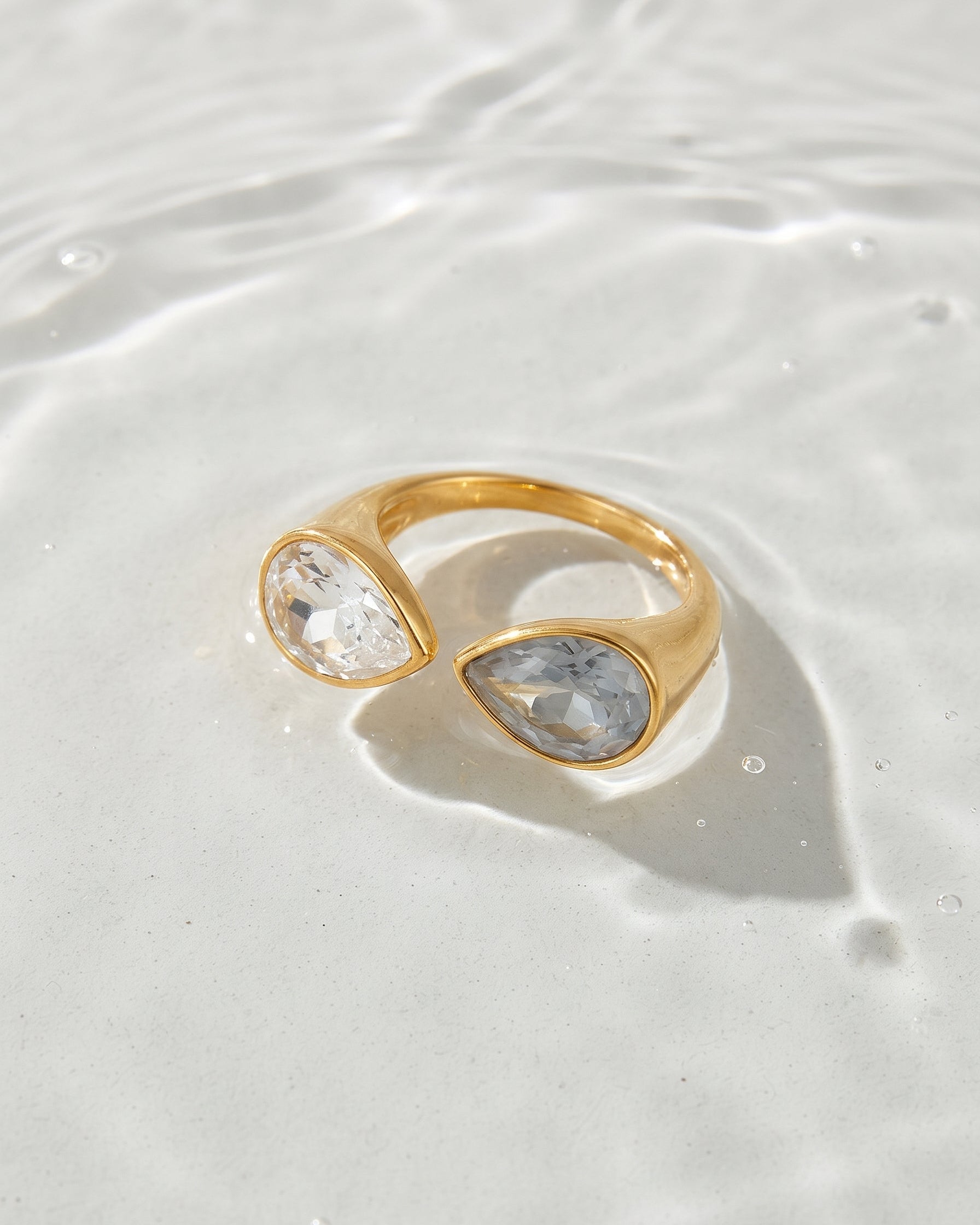 A gold ring with two teardrop-shaped clear gemstones, resting on a white textured surface with water ripples in the background.