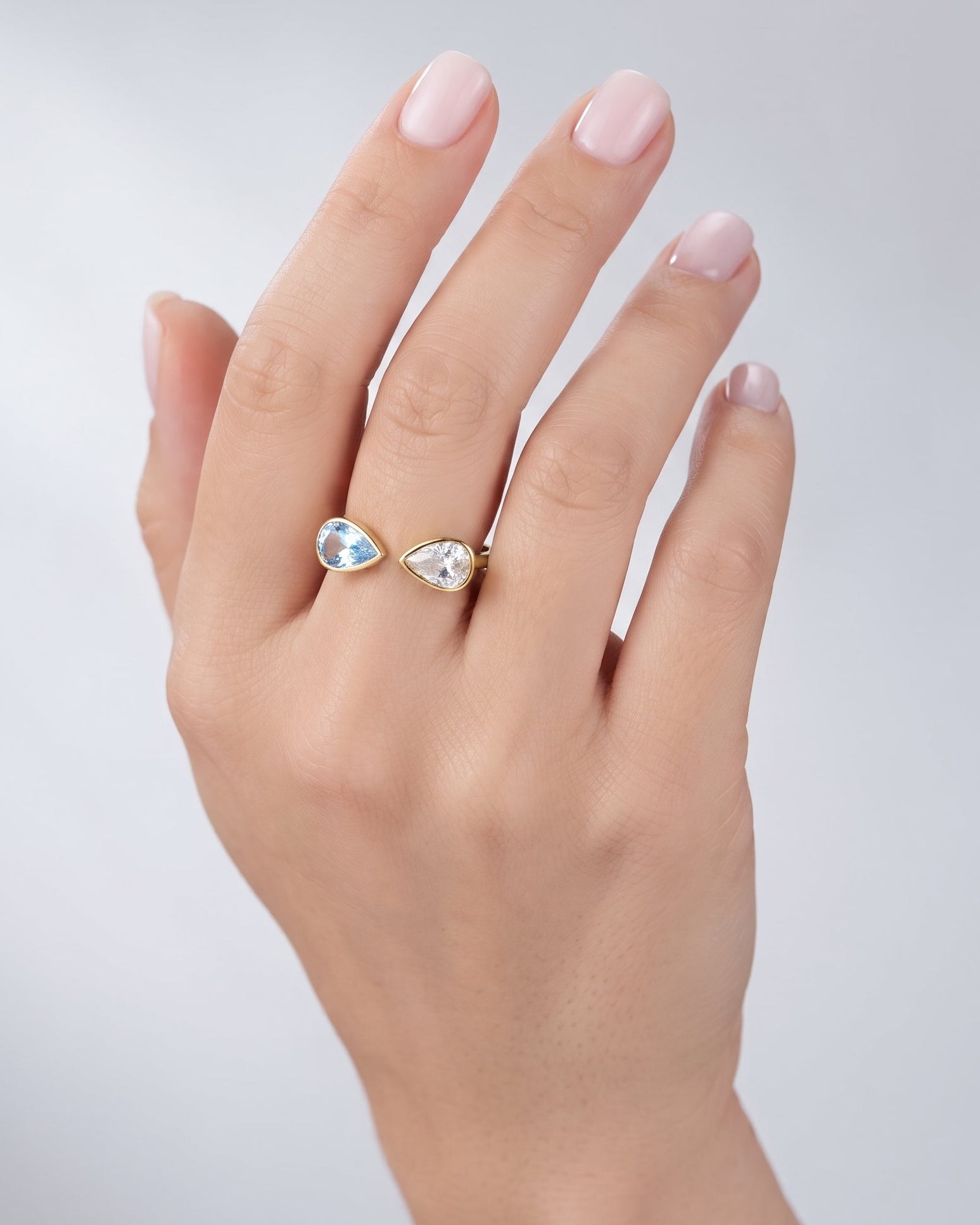 A close-up of a hand wearing a delicate gold ring set with two small pear-cut clear gemstones, photographed against a soft light gray background.