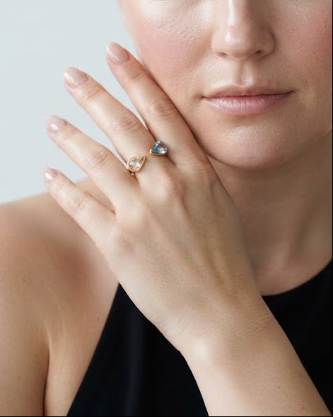 A close-up portrait of a woman with her hair pulled back, wearing a delicate gold ring with a small blue stone on one finger. She is wearing a black halter top against a light gray background.