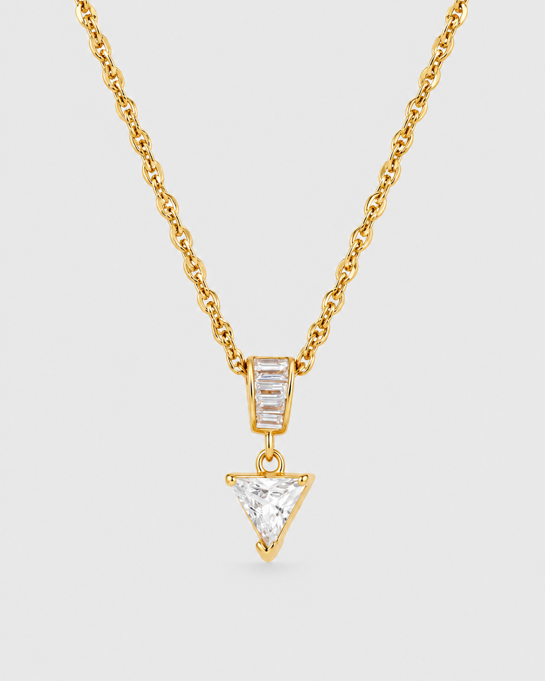 A gold chain necklace featuring a pendant with a baguette-cut clear stone and a dangling triangular clear stone, set against a light gray background.