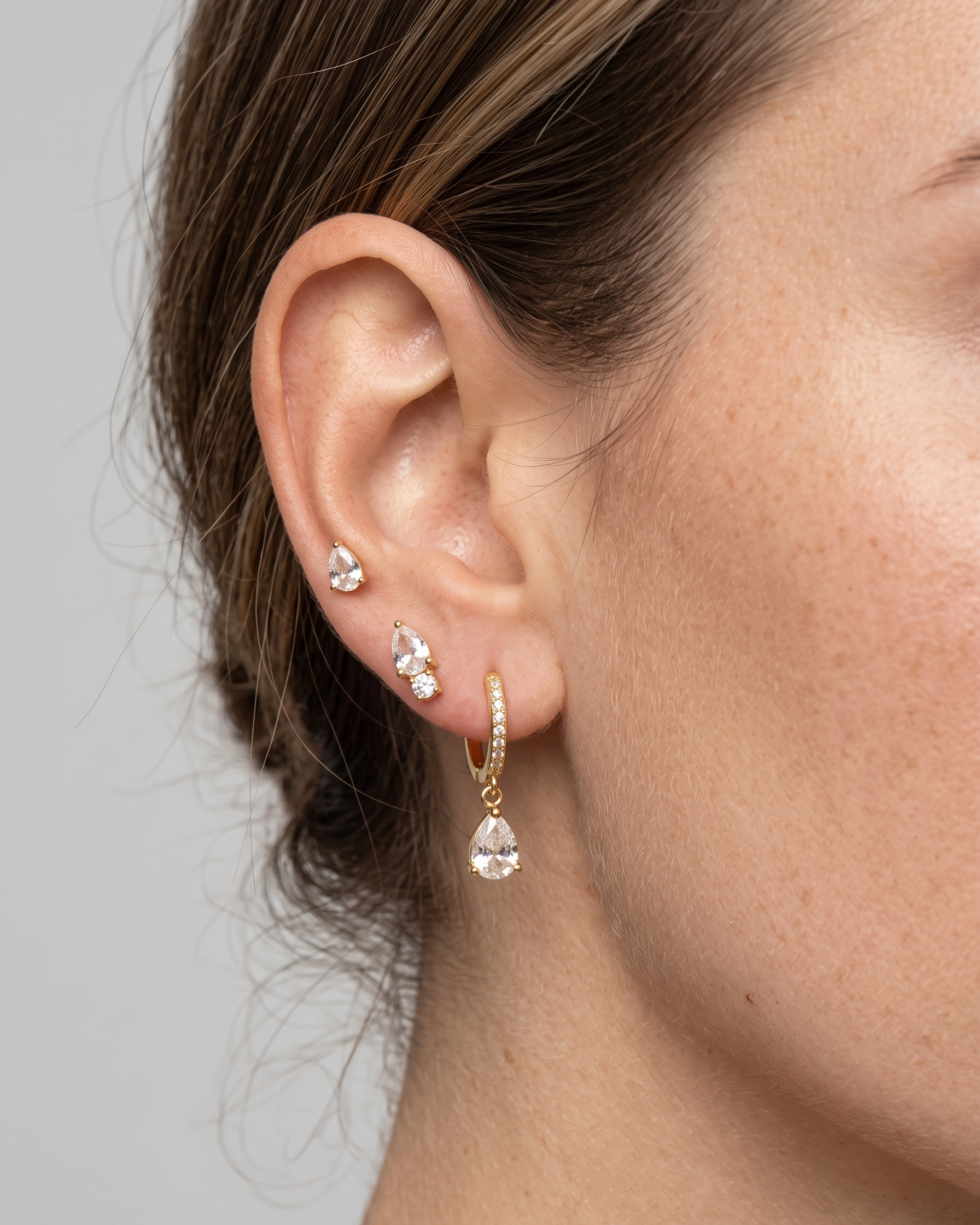Close-up of a person wearing gold earrings with clear gemstones against a neutral background