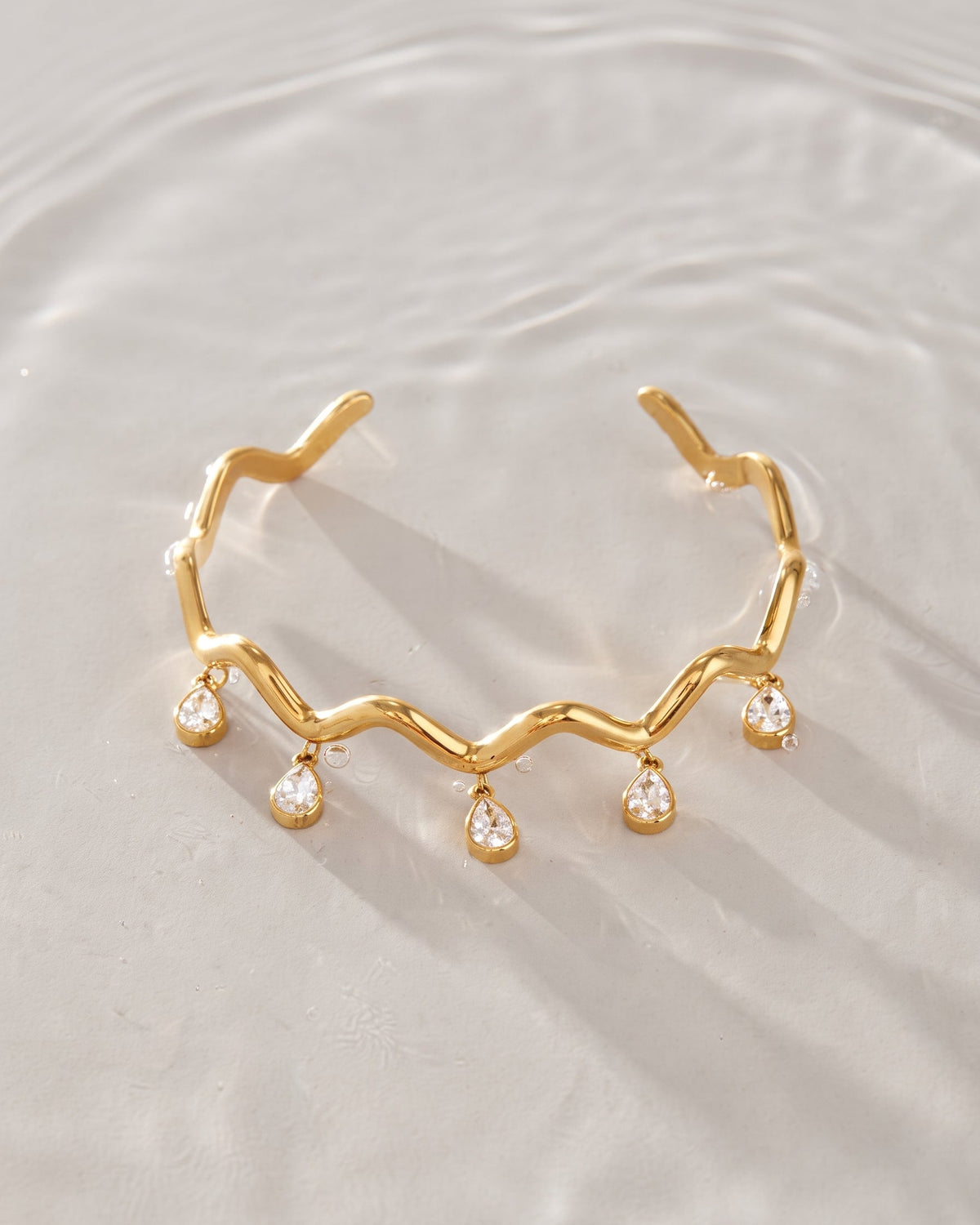 A gold open cuff bracelet with a wavy zigzag line and five dangling teardrop-shaped clear gemstones, photographed on a softly rippled light gray surface.