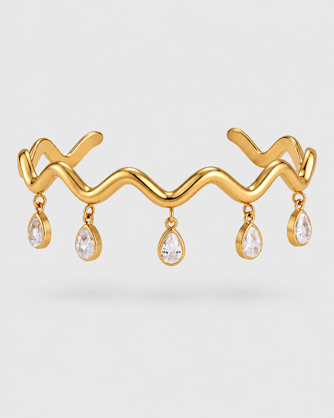 A gold open cuff bracelet featuring a wavy, zig-zag top design with five clear teardrop gemstones hanging from the front, set against a light gray background.