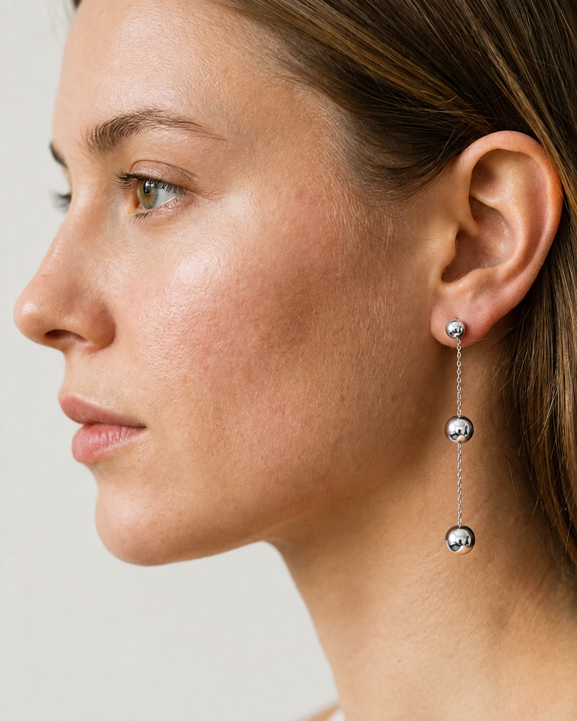 A close-up side profile of a woman wearing a delicate silver drop earring with three spherical beads suspended by thin chains.