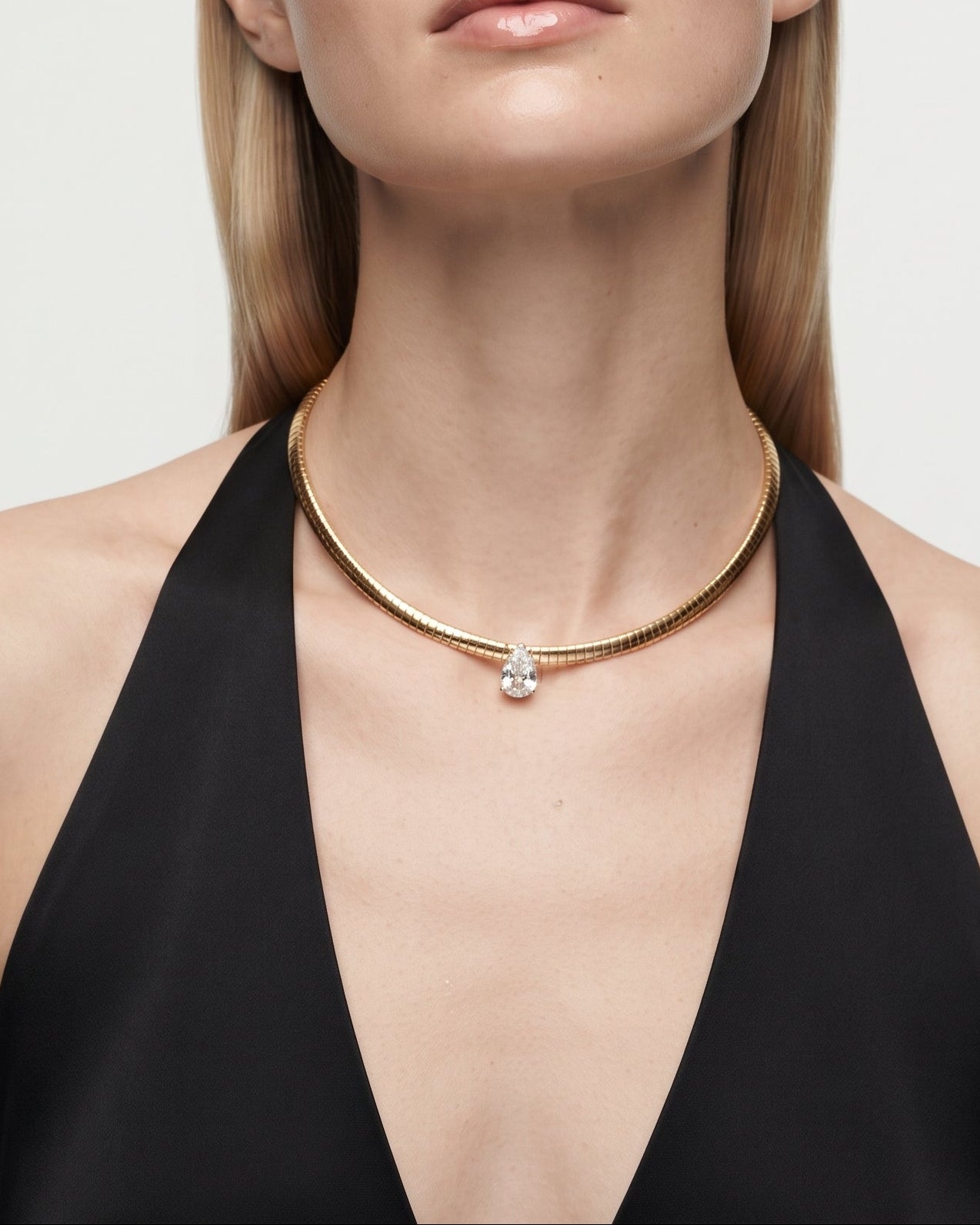 A blonde model wearing a delicate gold chain necklace with a small clear crystal pendant, styled with a black halter top against a light gray background.