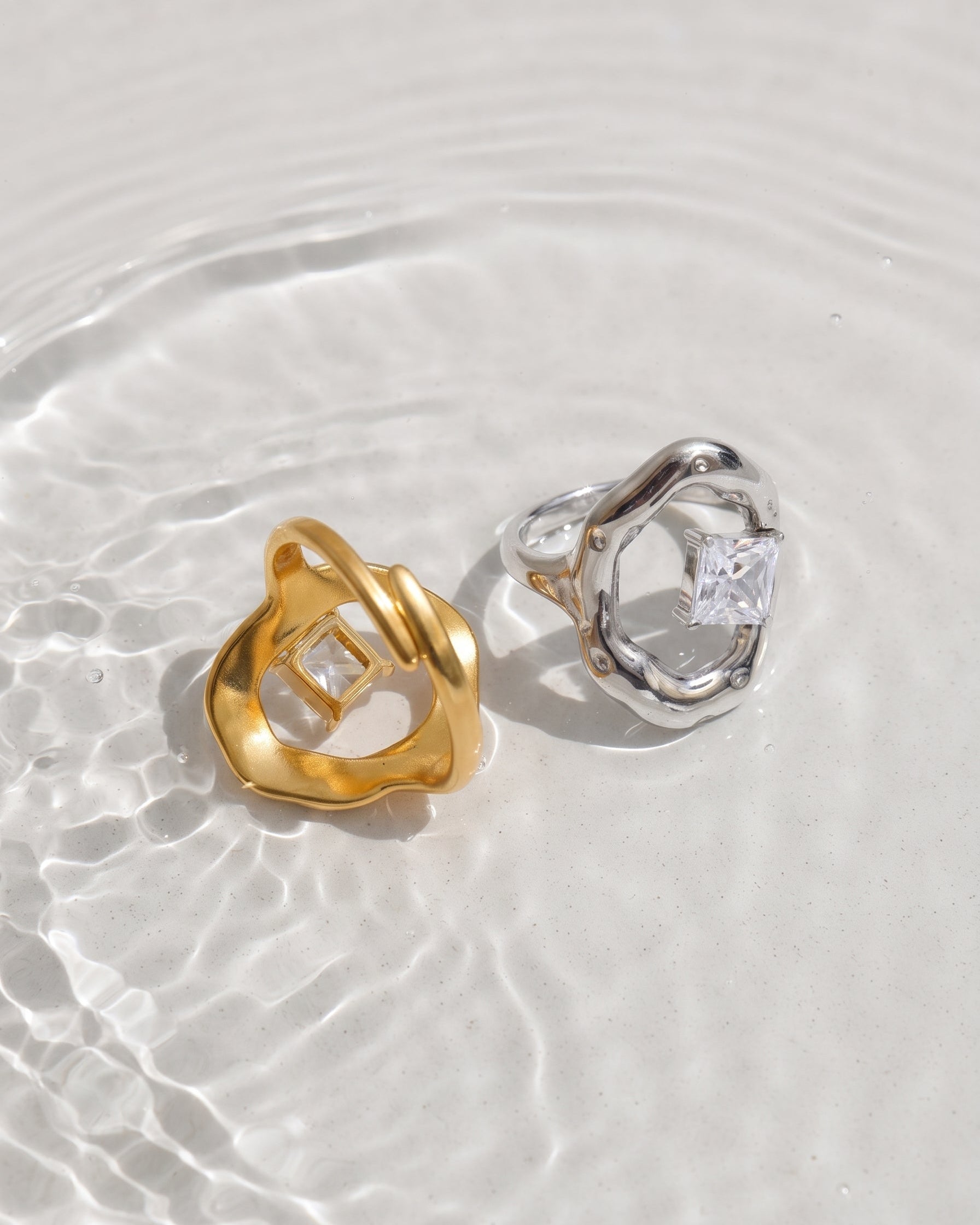 Two abstract sculptural rings, one in polished yellow gold and one in polished silver, each featuring a square clear gemstone, resting on a white textured surface with shallow water ripples.