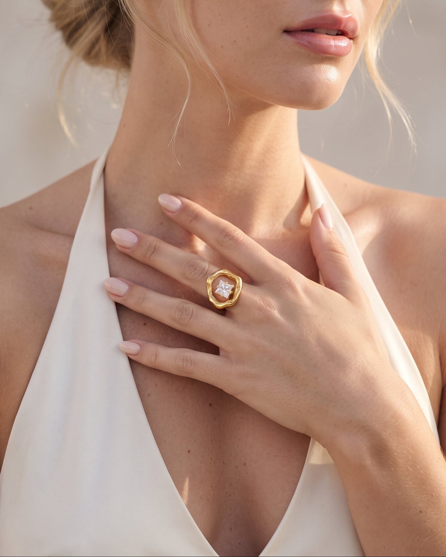 A close-up lifestyle shot of a woman wearing a gold ring with a clear central gemstone on her finger, paired with a cream halter top against a soft beige background.