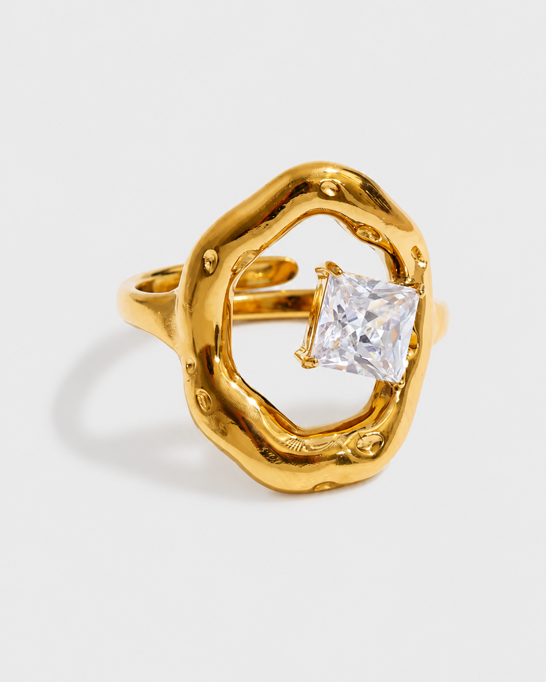 A gold ring with an organic, wavy openwork design and a central princess-cut clear gemstone, shown on a light gray background.